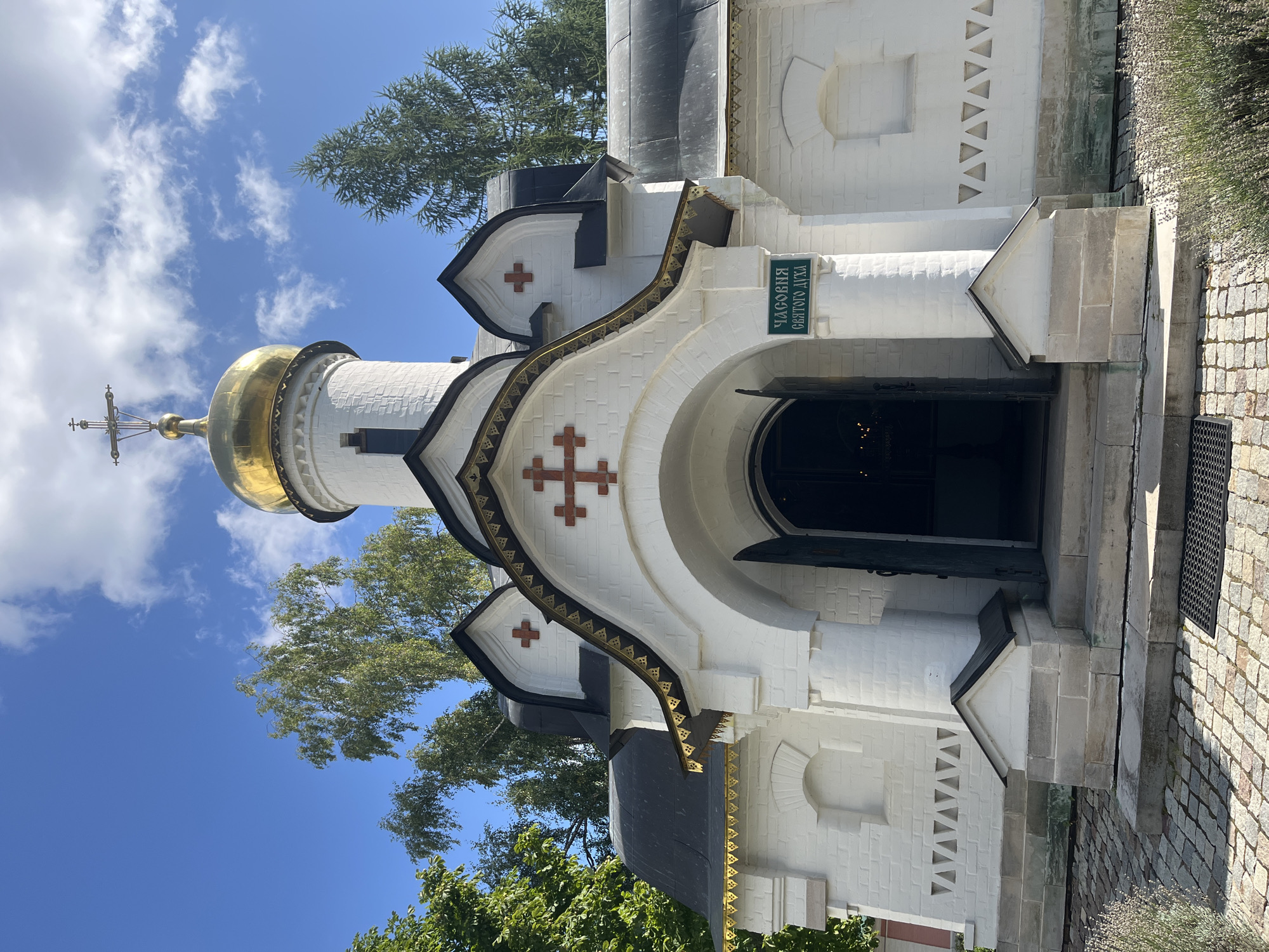 The image depicts the Chapel of the Holy Spirit in Dmitrov, Russia, on a clear, sunny day. The white brick chapel features a prominent golden dome topped with a cross. The entrance is an arched doorway with open doors, revealing the interior. Above the entrance, a green sign reads "ЧАСОВНЯ СВЯТОГО ДУХА" (Chapel of the Holy Spirit). Decorative red crosses are visible on the facade, and intricate gold trim adorns the rooflines. The chapel is surrounded by lush green trees, and the ground in front is paved with cobblestones. The sky is a vibrant blue with scattered white clouds. There are no people visible in the image.