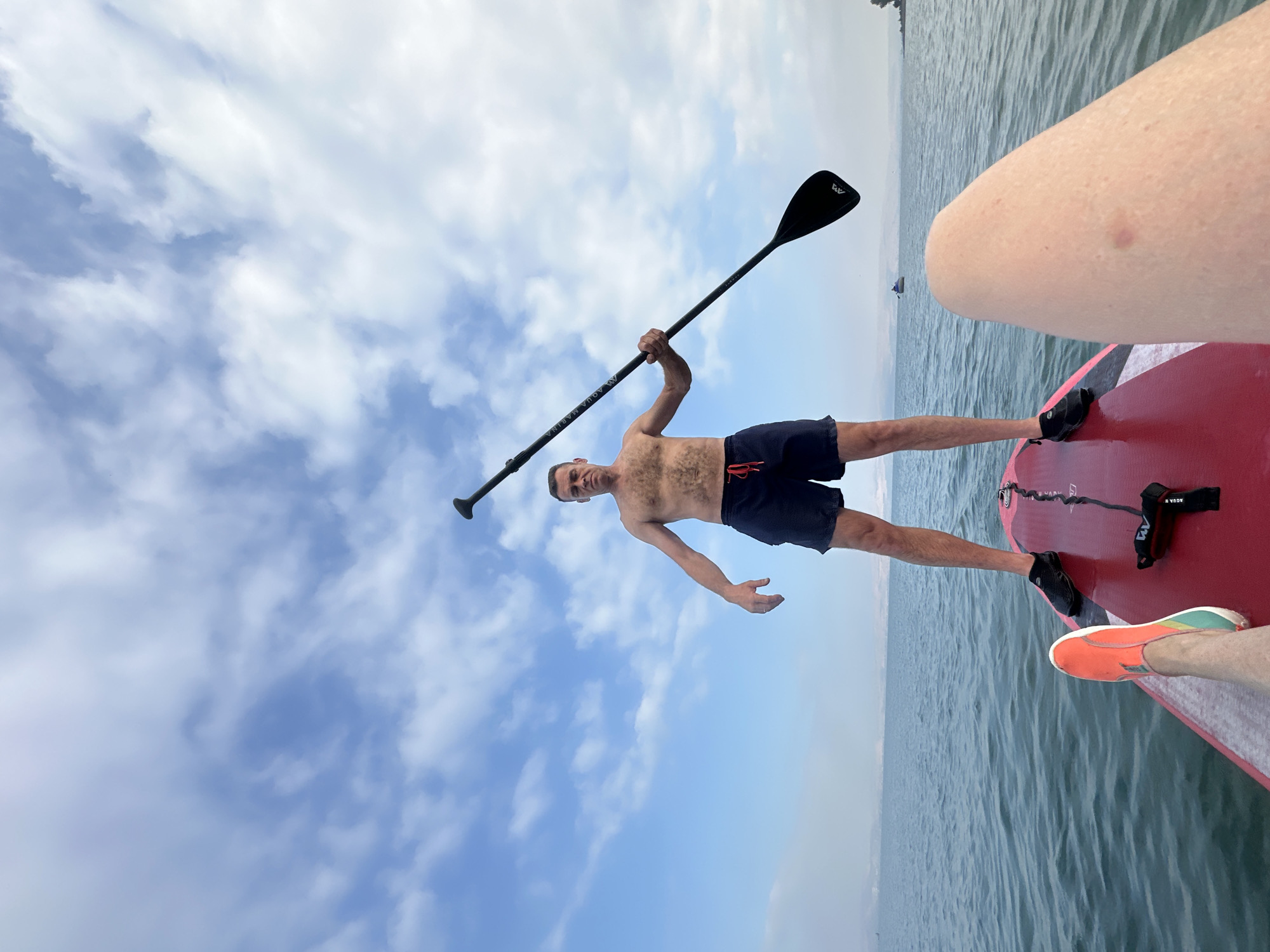 A man stands shirtless on a red paddleboard, holding a black paddle. He wears dark blue swim trunks with a red drawstring. His left arm is extended outward, while his right arm grips the paddle. The paddleboard is on calm, blue water under a sky with scattered white clouds. In the distance, land is visible along the horizon, suggesting the location is a body of water in Tiberias, Israel. The foreground shows another person's legs and feet, one wearing an orange and green shoe, resting on the paddleboard.