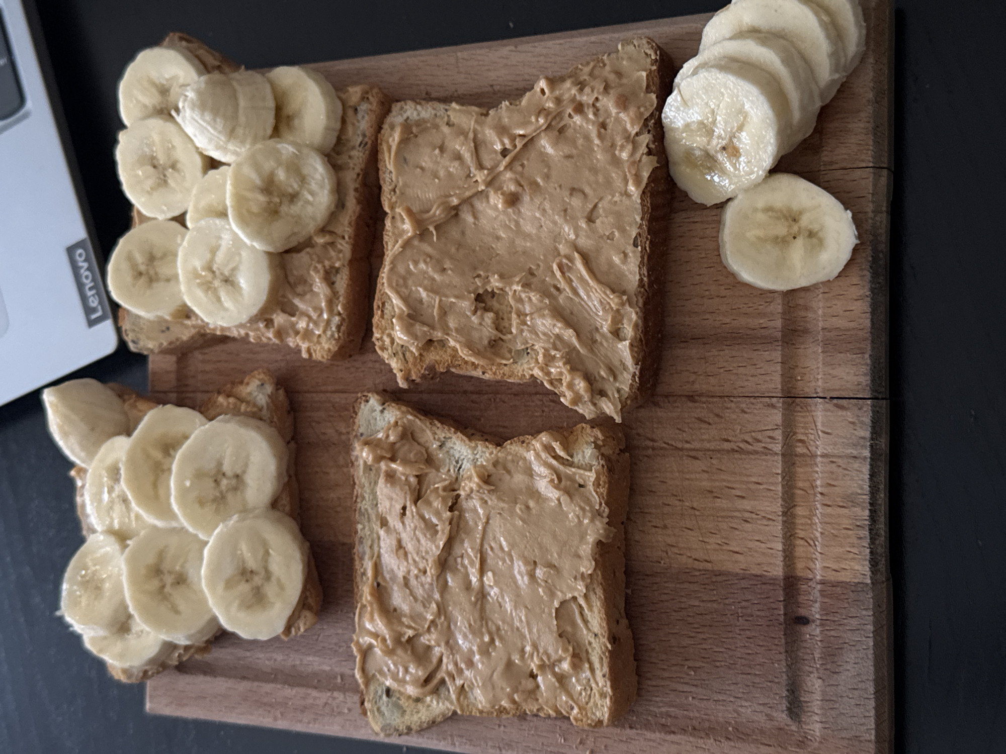 Four slices of toast are arranged on a wooden cutting board. Two slices on the left are spread with peanut butter, while the two slices on the right are topped with banana slices. A stack of sliced bananas is also on the cutting board to the right of the toast. A laptop is visible in the background. The scene appears to be a kitchen or dining area. The location is not specified.