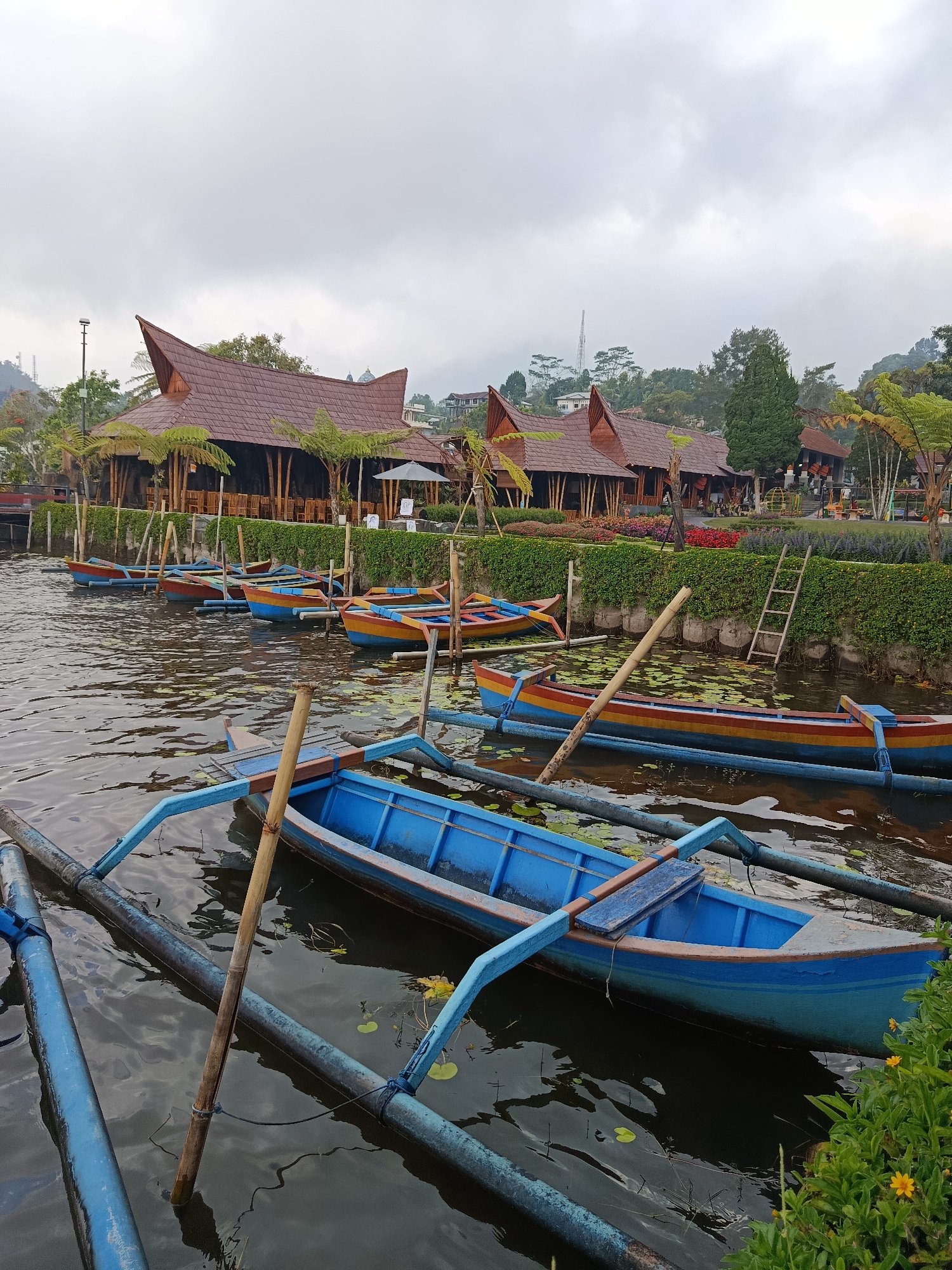A collection of brightly colored traditional Indonesian boats, some with outriggers, are docked on a body of water. The boats are primarily blue, red, and yellow. Several are tied to wooden posts in the water. In the background, there are several buildings with distinctive brown, pitched roofs, resembling traditional Balinese architecture. These structures are set against a backdrop of lush green vegetation and trees, with a hazy, overcast sky above. The location appears to be a scenic area in Baturiti, Indonesia, possibly a tourist attraction or a waterside village, given the presence of the traditional boats and buildings.