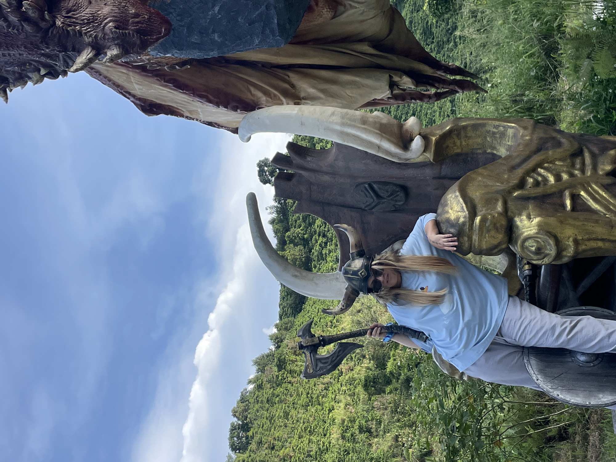 A woman, dressed as a Viking with a helmet and holding an axe, poses on a large, ornate throne-like sculpture. The throne is decorated with large horns and metallic gold accents. The woman is wearing a light blue t-shirt and grey sweatpants. Behind her, a large dragon statue is partially visible, with its wing and head in the frame. The scene is set outdoors, with lush green hills and a blue sky with white clouds in the background. This location is identified as Thôn 1, Vietnam.