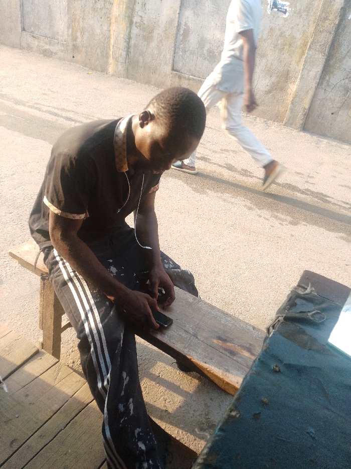 A man in a black polo shirt and black track pants sits on a weathered wooden bench, looking at his smartphone. He has earphones in. A second person, dressed in white, walks in the background on a paved surface. A dark-colored, box-shaped object with a shiny surface rests beside the man on the bench. The background is a light-colored building or wall. Sunlight illuminates the scene, casting shadows on the bench and surrounding areas.
