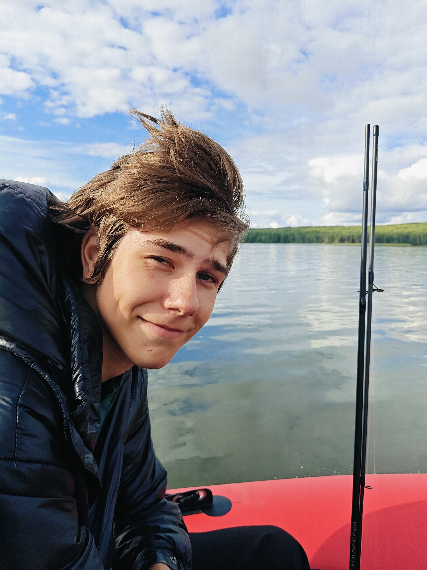 A young male, appearing to be in his late teens, is positioned in the foreground of the image, looking towards the camera with a slight smile. He has light brown hair that is windswept and he is wearing a dark, puffy jacket. The man is seated in a red inflatable boat, with a fishing rod visible to his right. The background depicts a calm body of water, likely a lake or river, with a treeline visible in the distance. The sky is blue with scattered white clouds. The setting appears to be outdoors in a natural environment. The image does not contain any elements that specifically place it within the city of Chaykovsky, Russia.