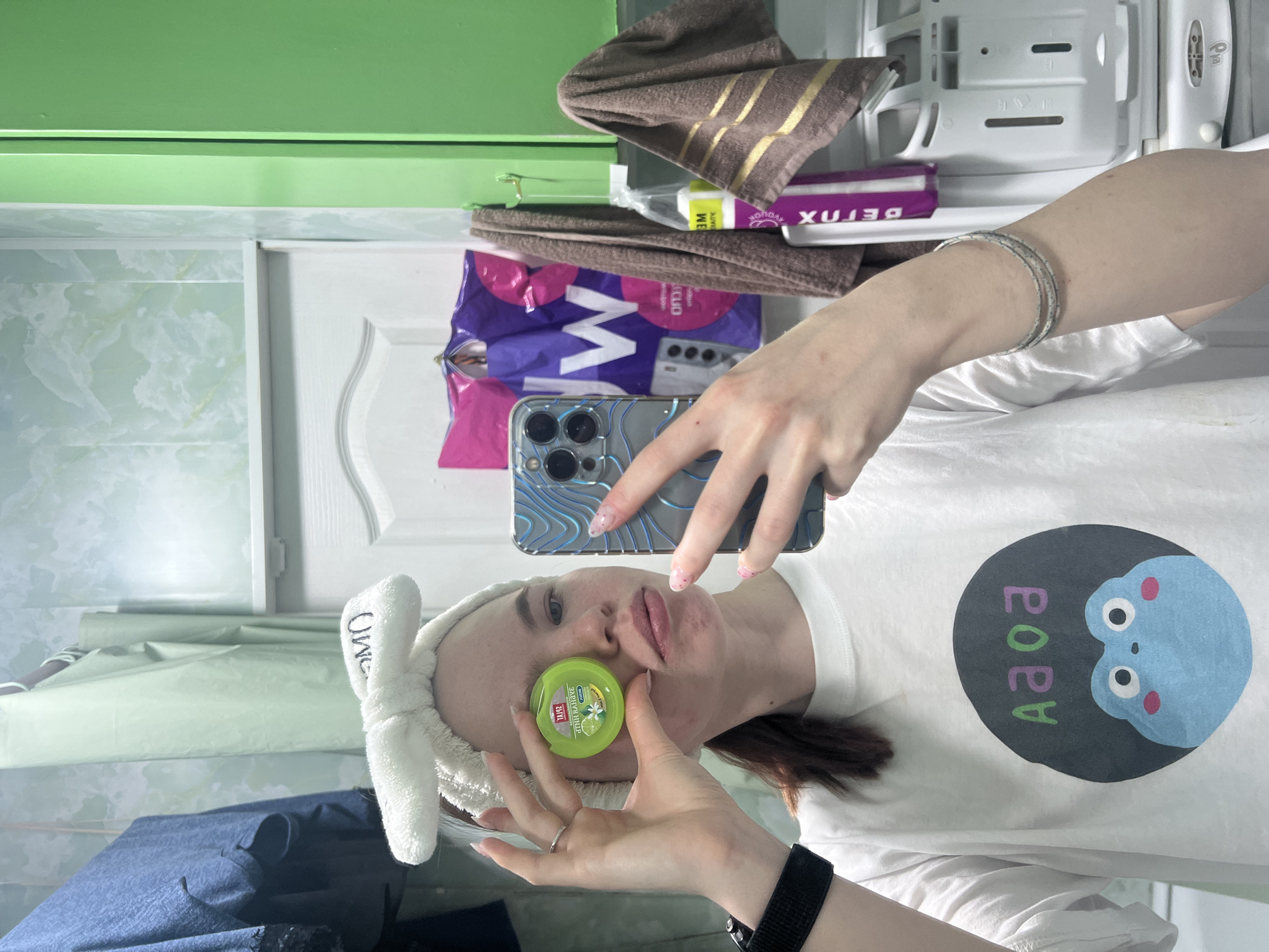 A rotated eye-level shot depicts a woman holding a small, round, green container in front of her face with both hands, appearing to take a selfie with a blue iPhone. She is wearing a white t-shirt with a cartoonish blue character on it and a white headband. The background shows what appears to be a bathroom setting with marble patterned wall tiles, a white door, and various toiletries and items on shelves. The location appears to be a bathroom in the city of Chaikovsky, Russia.