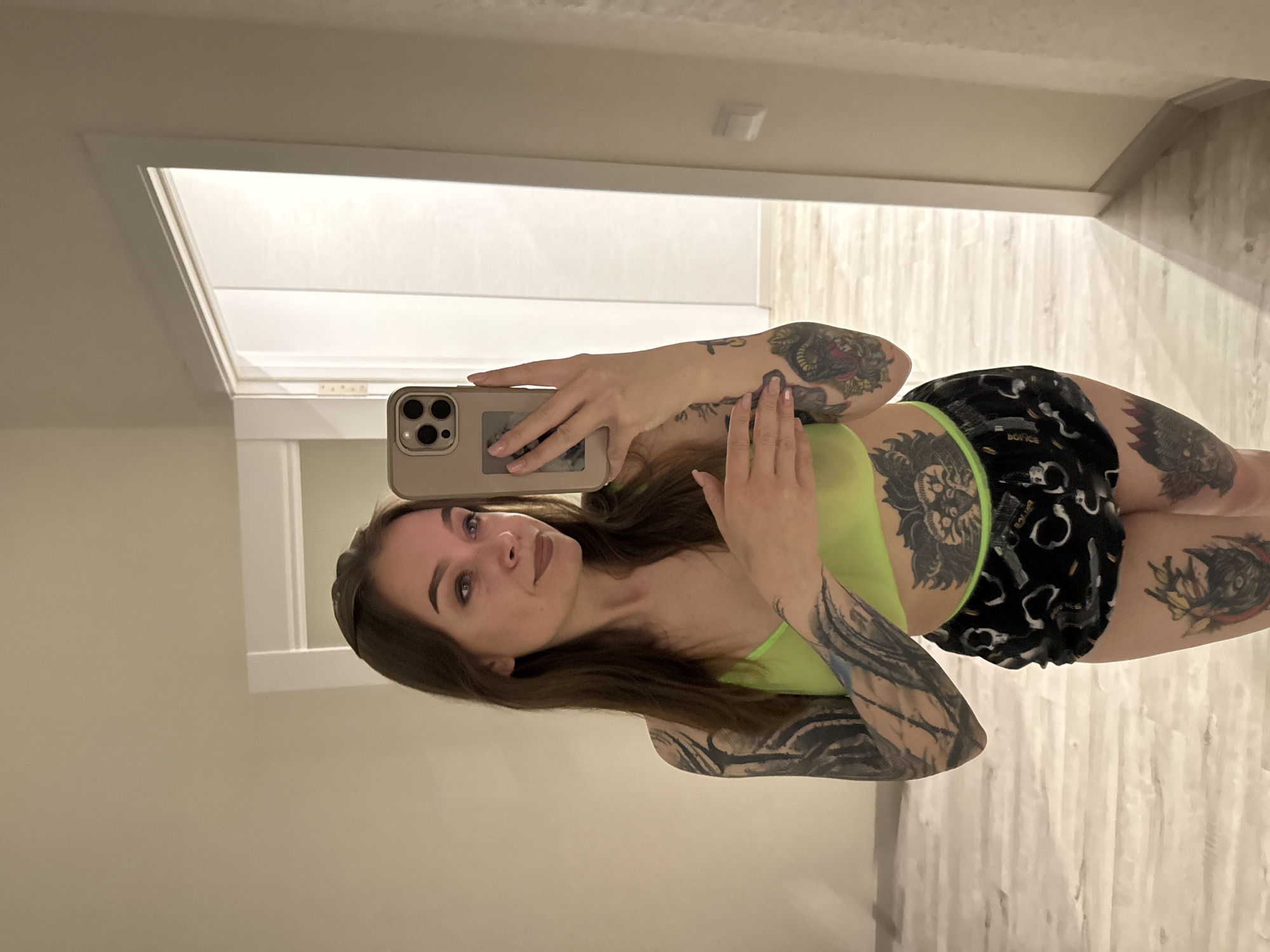 A young woman is depicted in a mirror selfie, captured indoors in what appears to be a modern residential or commercial setting in Gelendzhik, Russia. She has long, dark brown hair and a calm expression, looking directly towards the camera with a subtle smile. She is holding a light-colored smartphone, consistent with an iPhone model, in her right hand to take the photo of her reflection.
She is dressed in a neon green tank top or sports bra and dark shorts with a light, repeating pattern and a prominent bright green elastic waistband. Extensive tattoos adorn her body, including full sleeves on both arms with intricate designs and large figures on her thighs, notably what resembles a lion's head on her left thigh/hip area.
The background reflected in the mirror reveals light-colored walls and a light-toned wooden floor. White doorframes are visible, indicating an interior hallway or room. The word "BOYCE" is faintly discernible in white text on the upper right thigh of her shorts. The indoor lighting suggests a well-lit space, but the time of day or external weather conditions are not apparent.