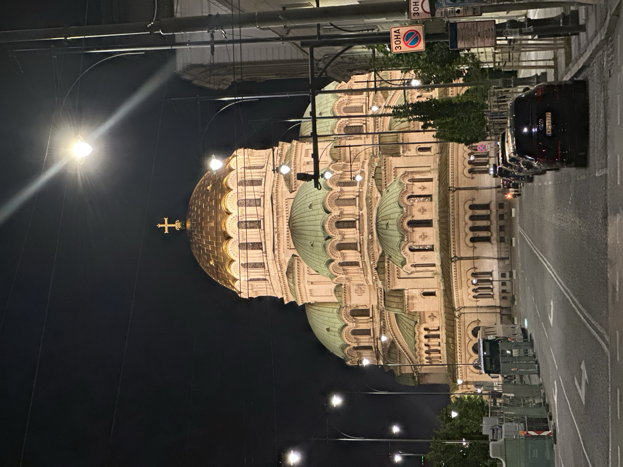 The image captures the illuminated St. Alexander Nevsky Cathedral in Sofia, Bulgaria, at night. The grand cathedral dominates the left side of the frame, showcasing its distinctive architecture with a large golden central dome topped by a cross, and several smaller, light green domes. The intricate pale stone facade of the building is highlighted by artificial lighting, making architectural details visible against the dark night sky.
On the right side of the image, a wide, empty street stretches into the distance, with white lane markings visible. A row of dark-colored cars is parked along the street. The license plate "HZ 7177" is discernable on one of the black cars. Numerous streetlights with bright, star-like glows and lens flares illuminate the street and foreground. Overhead power lines and utility poles crisscross the upper portion of the scene. Small trees with green foliage are visible along the street, breaking up the urban landscape.
A prominent street sign in the upper right displays "30HA" (ЗОНА), which means "ZONE," alongside a red circular sign with a blue background and a red diagonal line, indicating a "No Stopping" or "No Parking" restriction for that area. The time of day is clearly night, as evidenced by the dark sky and extensive artificial illumination of both the cathedral and the street. No people are visible in the scene, which conveys a serene and monumental atmosphere.