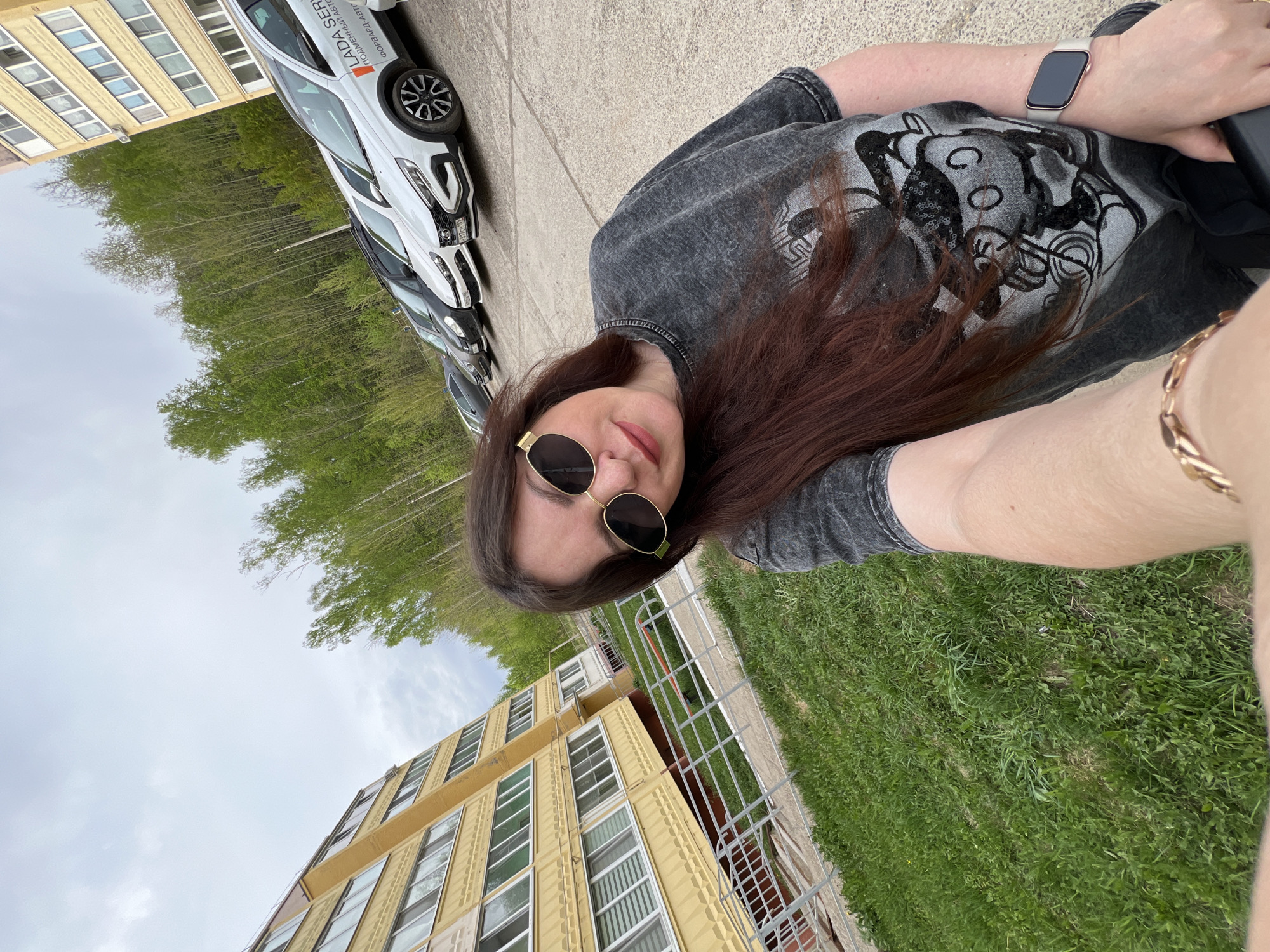 A woman wearing round sunglasses and a gray T-shirt with a graphic design is lying on grass near a building in Noviy, Russia. The building is yellow with multiple windows. A silver or white car is parked nearby. The woman has brown hair and is wearing a watch and a bracelet. Trees are also visible in the background.