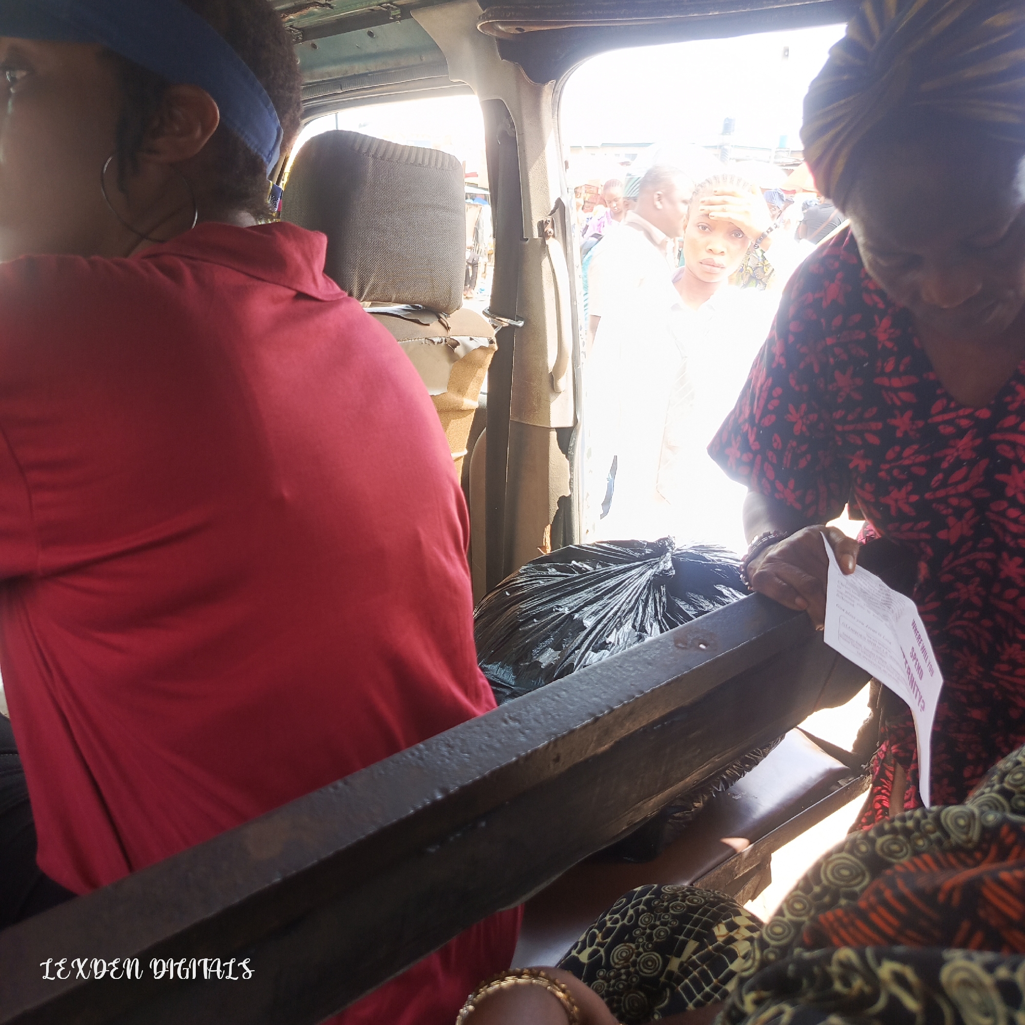 In this image, three individuals are visible inside a vehicle, possibly a bus or van, in Egan, Nigeria. The foreground is dominated by a person on the left wearing a red shirt and a blue headband. To their right, a black trash bag is partially visible. In the middle ground, a person in a white shirt is seen with their hand on their forehead, looking distressed. Another person, wearing a black and red patterned dress, is in the right foreground, holding a small leaflet. The background depicts a bright, outdoor scene, suggesting it is daytime with sunny weather. The overall atmosphere seems slightly chaotic, with people visible outside the vehicle, hinting at a busy market or roadside setting. The leaflet held by the woman in the patterned dress has visible text in English, including the question "WHERE WILL YOU SPEND ETERNITY?". The logo "LEXDEN DIGITALS" is watermarked at the bottom left of the image. The interior of the vehicle appears worn, with damaged seat coverings.