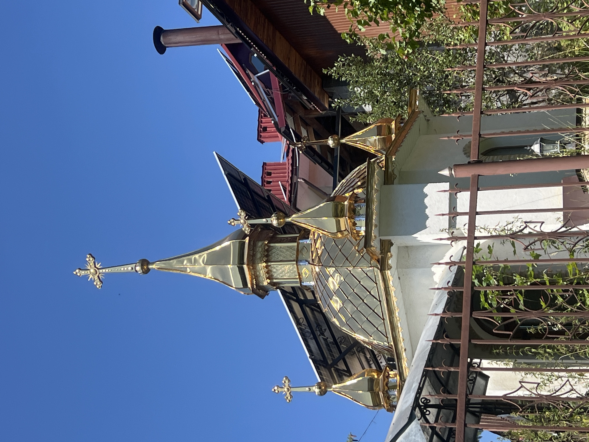 The image shows the golden domes of a small orthodox church or chapel against a clear blue sky in Yaremche, Ukraine. The church has multiple ornate golden domes, each topped with a cross. The main dome is larger and more elaborately decorated, with a translucent material on its upper section. Smaller, pointed golden domes surround the main one. The building itself is white with golden trim. In front of the church, there is a rusty metal fence with some greenery growing through it. Behind the church, the roofs of other buildings are visible, including red tiled roofs and some with solar panels. Trees and foliage are also present in the background. The scene is bathed in bright sunlight, suggesting a clear day.