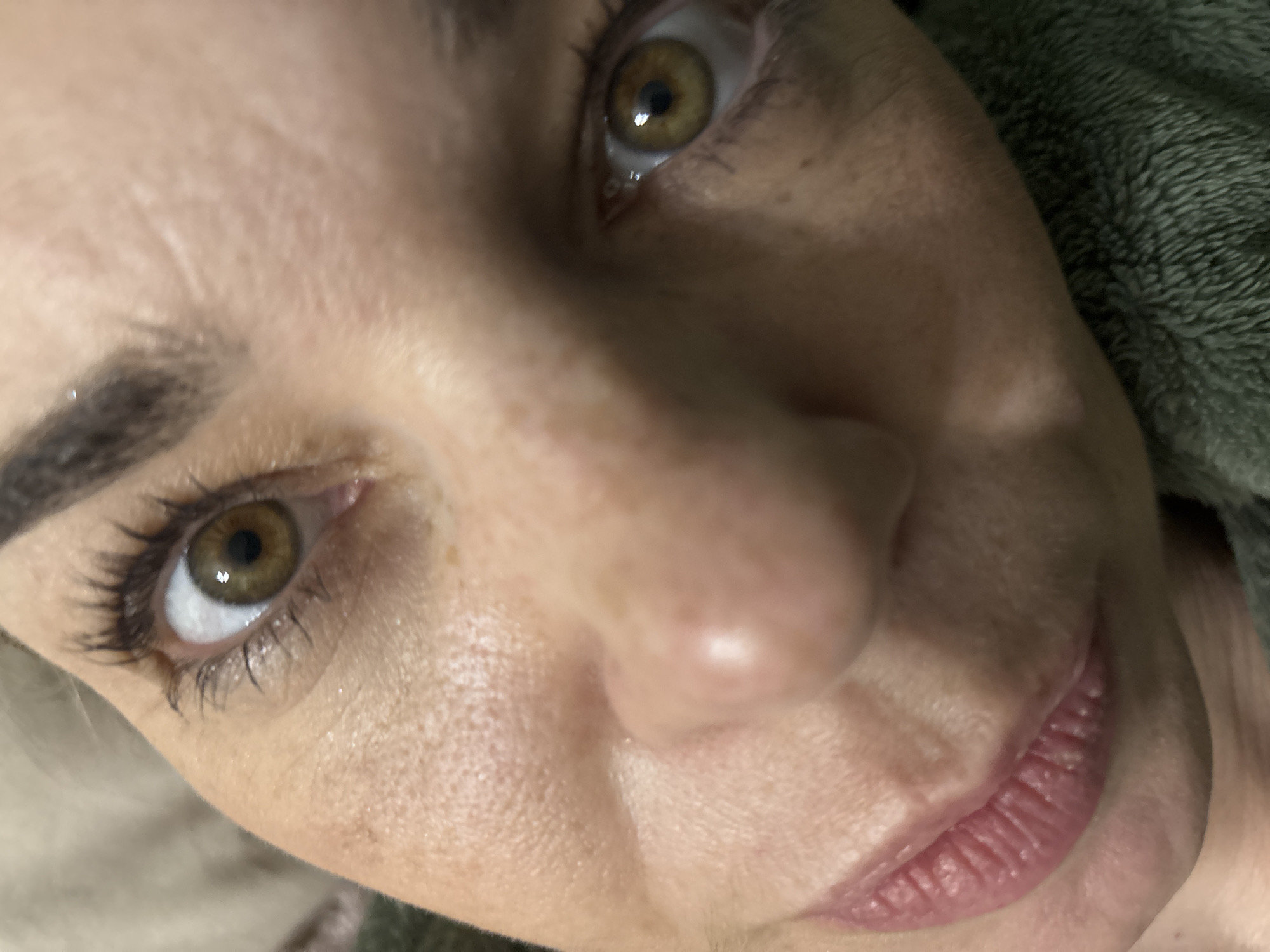 A close-up, angled image focuses on a woman's face. She has fair skin with visible pores and some fine lines. Her eyes are hazel, with dark eyelashes and eyebrows. The right eye appears to have a slight tear. Her lips are pink and slightly parted. The background includes a soft, green fabric texture, possibly a towel or blanket. The image contains no contextual information to indicate a location in Vsevolozhsk, Russia.