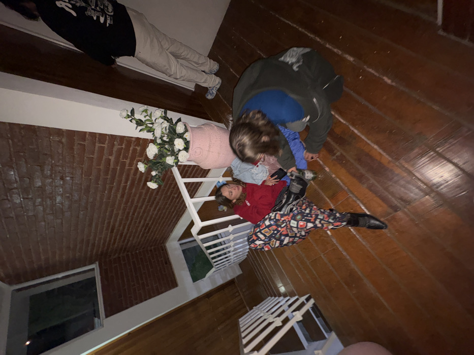 A group of people is positioned on a staircase. One person is lying down on the stairs wearing a red sweatshirt and patterned pants. Another person is leaning over them, and a third individual is standing nearby. A large pink vase with white flowers sits next to the staircase. A fourth person is standing by the top of the stair, wearing dark clothing and cream-colored pants. The setting appears to be indoors with wooden floors and a brick wall. A window is visible in the background.