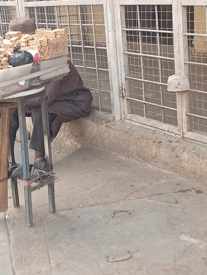 In Gawuna, Nigeria, an outdoor scene depicts a street vendor operating near a building. A person, partially visible and seated, wears a dark brown garment and a brown cap, with their feet resting on the lower crossbar of a simple vendor's table. The table features an unpainted wooden top supported by light blue/grey painted metal legs. On the table, multiple stacks of light brown, rectangular items (likely food products) are displayed for sale. A black plastic bag and a small red object are also on the table. Behind the vendor, a building facade with white or light-colored metal grates covers window or door openings, forming a grid pattern. A raised concrete ledge runs along the base of this structure. Attached to the right-side vertical grate frame is a white, helmet-like object with two dark circular markings. The ground is a light grey concrete surface with minor imperfections. Two U-shaped metal handles are embedded in the concrete in the foreground. The scene is illuminated by natural light.