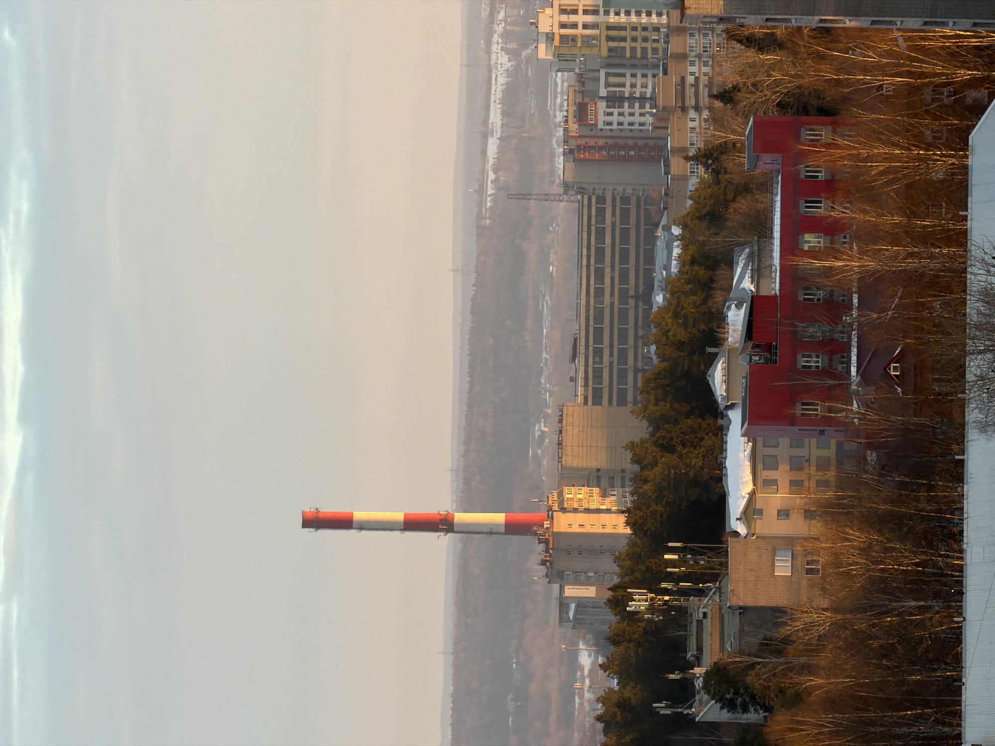 An elevated, long-distance forensic view captures an urban and semi-industrial landscape in Izhevsk, Russia, likely during winter, illuminated by the low-angle, warm light of either sunrise or sunset.
The most prominent feature is a tall industrial chimney, centrally located on the left, displaying alternating red and off-white/yellowish horizontal stripes along its entire visible length. Ladder-like structures are affixed to its sides. This chimney is characteristic of the Izhevsk Mechanical Plant (Izhmekh) / Kalashnikov Concern industrial complex, situated in the northern part of central Izhevsk, near Promyshlennaya Street.
Below and surrounding the chimney, a mix of multi-story buildings is visible. These include a concrete-colored building directly at the chimney's base, a large, horizontally oriented administrative or industrial structure with multiple window bands to its right, and various residential-style buildings of differing heights and architectural styles, some with snow-dusted rooftops. In the lower-midground, a distinctive red multi-story building with numerous windows stands out amidst other structures and bare deciduous trees.
Interspersed throughout the midground are dense clusters of trees, including darker evergreen species and numerous bare deciduous trees, their branches illuminated by the warm sunlight. Several rooftops feature visible cellular communication towers with antenna arrays.
The background consists of a vast, hazy, low-lying forested area, appearing bluish-grey, with patches of white indicating snow cover. Faint, distant structures, possibly power line towers or smaller industrial elements, are discernible against the horizon.
The sky transitions from a pale blue-grey at the top to a faint pinkish-orange gradient near the horizon, indicative of a low sun angle. The overall lighting suggests a clear, possibly cold, atmosphere. No discernible human figures, vehicles, or specific activities are visible within the frame.
