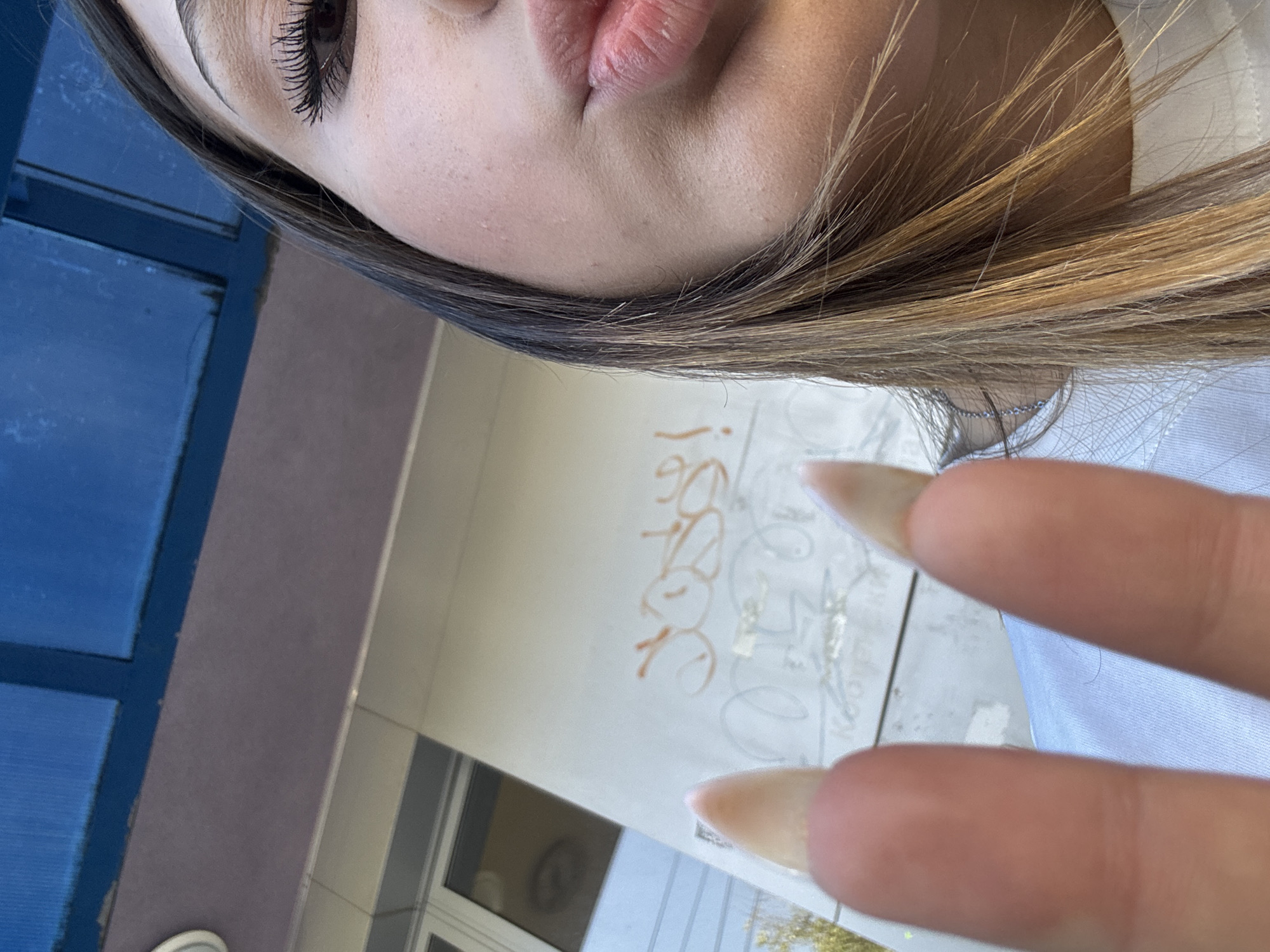 A close-up, eye-level shot captures a young woman's face and hands. She is outdoors, with a building and graffiti visible in the background, as well as a blue overhead structure. The woman has long, light brown hair with blonde highlights and is wearing a white shirt. Her right eye is visible, with mascara on her lashes. Her lips are slightly parted. She is holding up two fingers on her left hand, which has long, almond-shaped nails. The background is somewhat out of focus, with graffiti appearing on the white wall. The lighting suggests it is daytime.