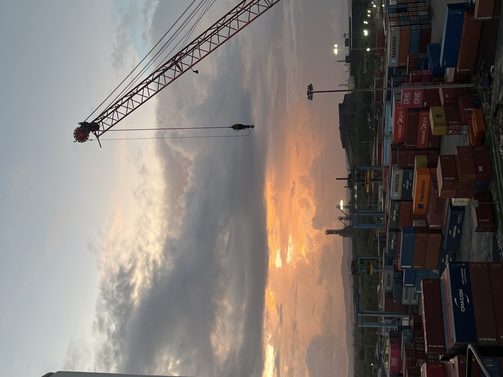 The image depicts a port facility at dusk, with a dramatic sky split between grey clouds and an orange sunset. A red crane dominates the left side of the frame, its boom extending towards the sky. On the right side, stacks of shipping containers, many with company logos such as "MAERSK," "ONE," "HAMBURG SÜD," and "CMA CGM," fill the frame. Streetlights are visible in the background, suggesting an urban or industrial setting. The image is taken from a high vantage point, looking down on the port.