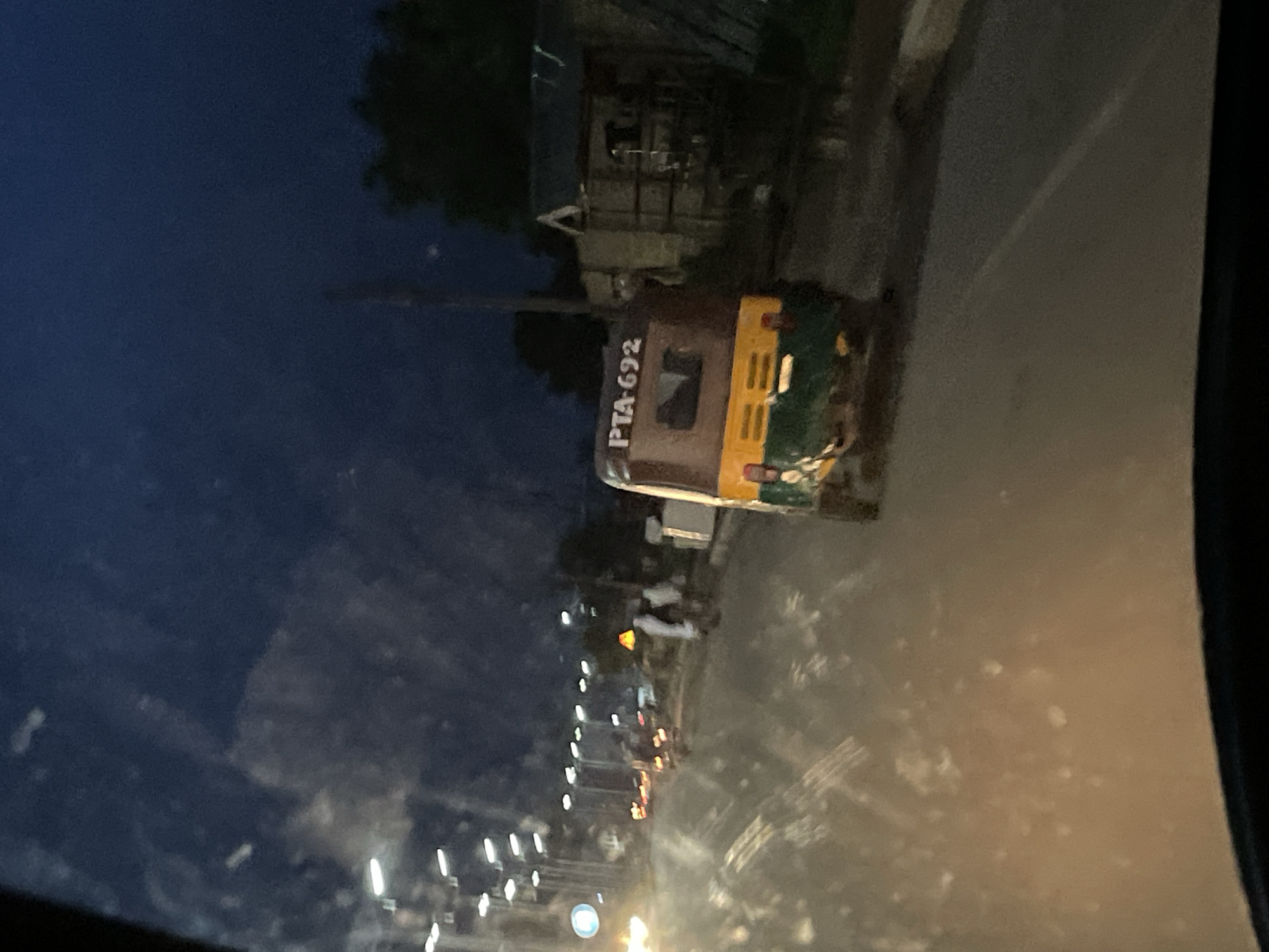 The image depicts a nighttime street scene in Jos, Nigeria. A three-wheeled auto-rickshaw, identified by the license plate "PTA-692", is prominently featured in the foreground, traveling on a paved road. The auto-rickshaw has a brown roof and cab, with a yellow and green lower body. Streetlights are visible in the background, casting a glow on the wet road, suggesting recent rainfall. Buildings and trees are dimly visible in the distance. The overall atmosphere is dark and hazy, typical of urban environments at night.