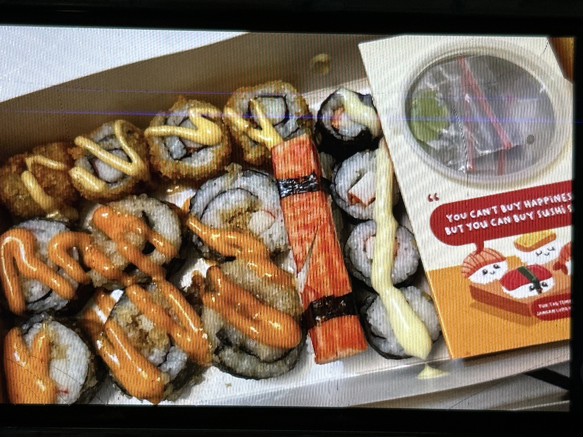 The image shows a close-up, overhead view of a takeout sushi box filled with various sushi rolls. The sushi appears to be a mix of styles, with some rolls topped with orange sauce, others with a creamy yellow sauce, and some covered in crispy fried bits. Several rolls are wrapped in seaweed and rice, with visible fillings. One section of the box contains what appears to be imitation crab sticks.
A small, orange, folded card is visible on the left side of the box, featuring cartoonish illustrations of sushi characters and text in red that reads, "YOU CAN'T BUY HAPPINESS BUT YOU CAN BUY SUSHI." Below this, smaller text is present, possibly a brand name or slogan.
To the right of the card, a circular, clear plastic container with a lid is placed inside the box. The container appears to hold some green leaves, possibly garnishes or accompanying ingredients, although the contents are somewhat obscured.
The overall environment is a white, textured surface, suggesting a table or a similar flat surface, typical of a dining or food preparation area. The lighting is somewhat diffused, and there are some vertical lines of light and color distortion across the image, indicating the photo was taken on a screen or with a camera experiencing interference.
While the image itself does not contain explicit geographical markers, the context of a takeout sushi box with promotional material and the mention of "Depok, Indonesia" implies that this scene likely takes place within a food establishment, restaurant, or a residential setting in Depok, Indonesia, where such food items are commonly purchased and consumed.