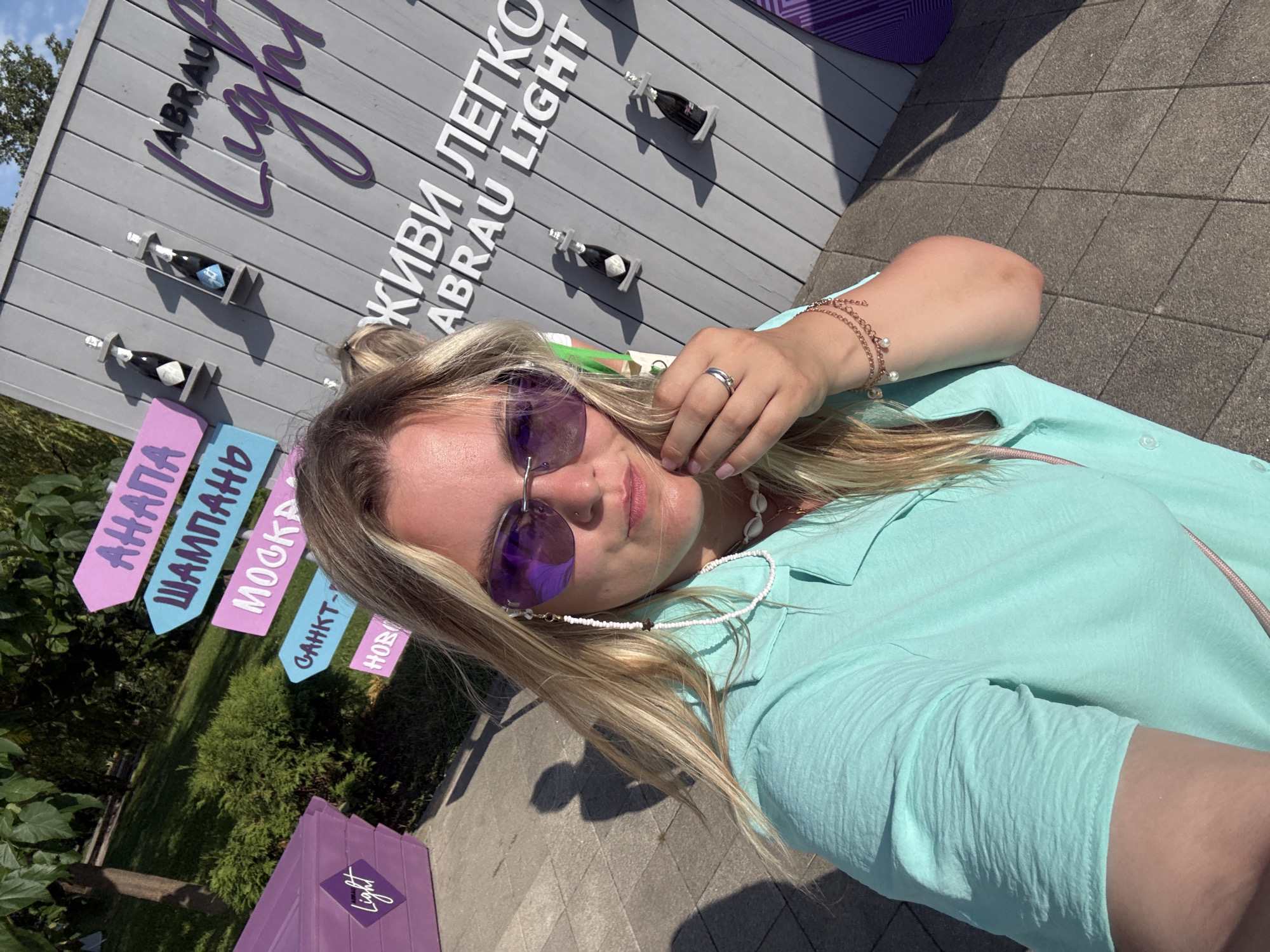 A blonde woman with sunglasses and a light blue shirt is taking a selfie in front of a sign in Abrau-Dyurso, Russia. The sign has purple lettering that reads "ABRAU Light" and "ЖИВИ ЛЕГКО ABRAU LIGHT." There are also several small bottles of what appears to be champagne displayed on the sign. To the left of the sign, there is a purple structure with the word "Light" written on it, and a directional sign with several destinations listed, including "Анапа" (Anapa), "Шампань" (Champagne), "Москва" (Moscow), "Санкт-" (Saint-), and "Ново" (Novo). The ground is paved with grey tiles. The background shows trees and a clear blue sky.