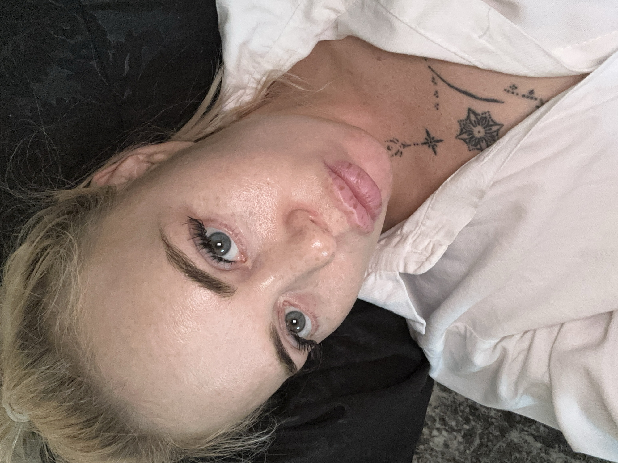 A close-up, high-angle selfie captures a fair-skinned woman with blonde hair pulled up in a messy bun. Her eyes, with visible eyelashes and light blue irises, gaze forward. Her lips are slightly parted, revealing a hint of teeth. She wears a white collared shirt, open at the neck, revealing a small tattoo on her sternum. The background is dark and out of focus, with a textured surface visible in the lower left corner.