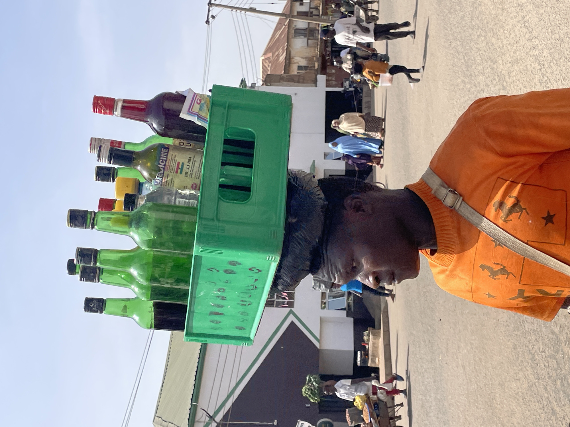 A portrait-oriented image captures a street scene, likely in Bukuru, Nigeria, focusing on a person carrying goods on their head. The main subject, seen from the mid-torso up and facing right, has dark skin and wears a bright orange long-sleeved shirt patterned with horses and stars. A light-colored strap crosses their left shoulder.
Resting on their head, stabilized by what appears to be a black cloth or padding, is a green plastic crate. The crate is filled with various bottles. Some are empty green glass bottles, while others contain liquids of different colors, including dark red, dark green, and yellow. One green bottle prominently displays a label with "MEDICINE" and "DR. JJ JIBA" along with what looks like a Nigerian flag graphic (green, white, green). Another bottle on the top right has a partial label starting with "G...". The crate and bottles are balanced skillfully on the head.
In the blurred background, several other people are visible on a paved street. Some figures are walking, including individuals dressed in a blue hijab/veil and patterned skirts. Buildings with varying architectural styles, including one with a corrugated iron roof, line the street. The bright, clear sky and strong sunlight suggest it is daytime, casting visible shadows on the ground. The overall atmosphere depicts a bustling daily life scene.