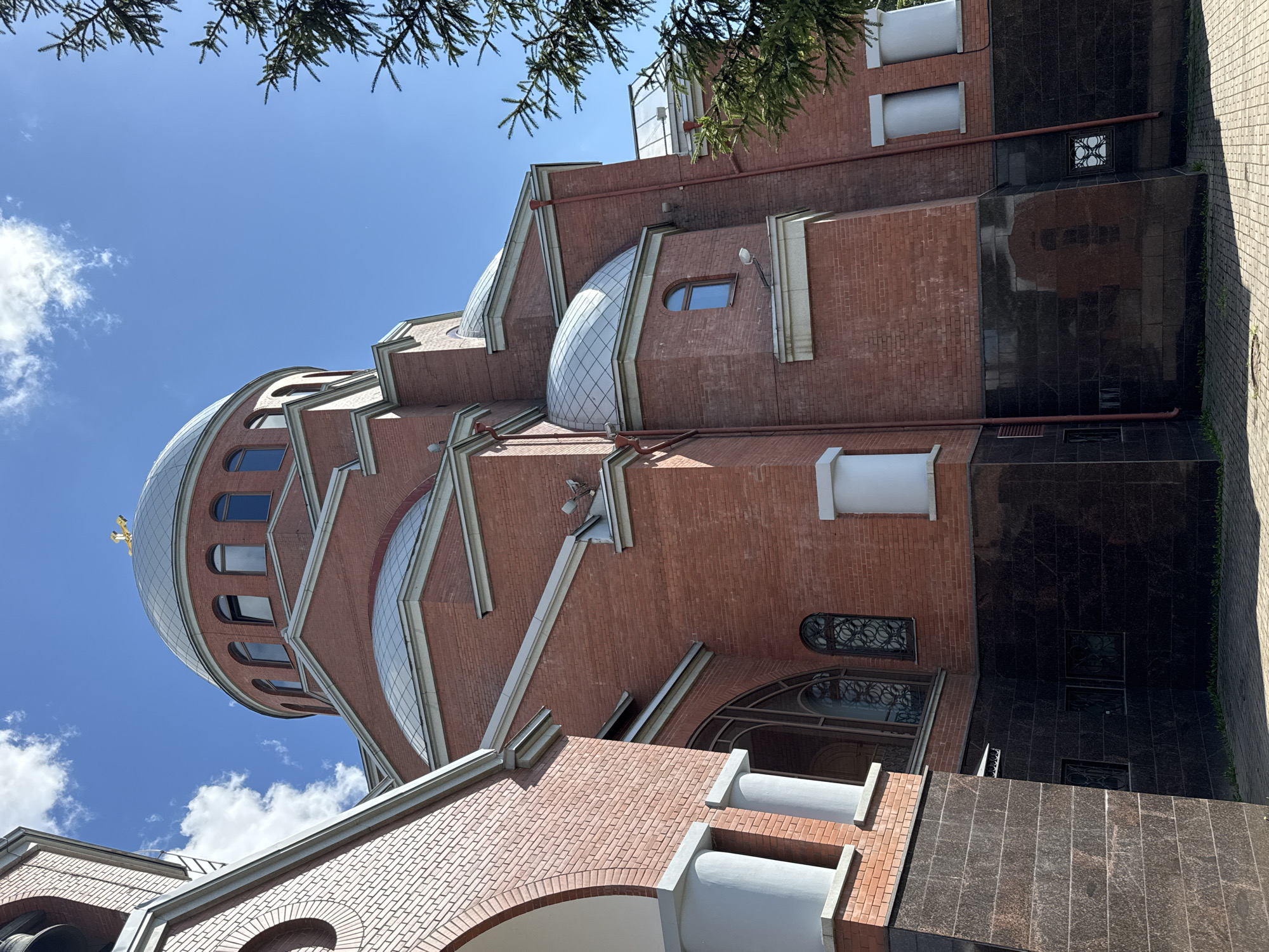 The image captures the exterior of a red brick church with multiple domes, located in Saint Petersburg, Russia. The architectural style suggests an Orthodox Christian church, characterized by its onion domes and a golden cross topping the main dome. The building features arched windows with ornate grills and contrasting darker stone at the base. The sky is clear blue with scattered white clouds. A section of a pine tree branch is visible on the right side of the frame, suggesting an outdoor setting with vegetation. The ground is paved with cobblestones. There are no people visible in the image.