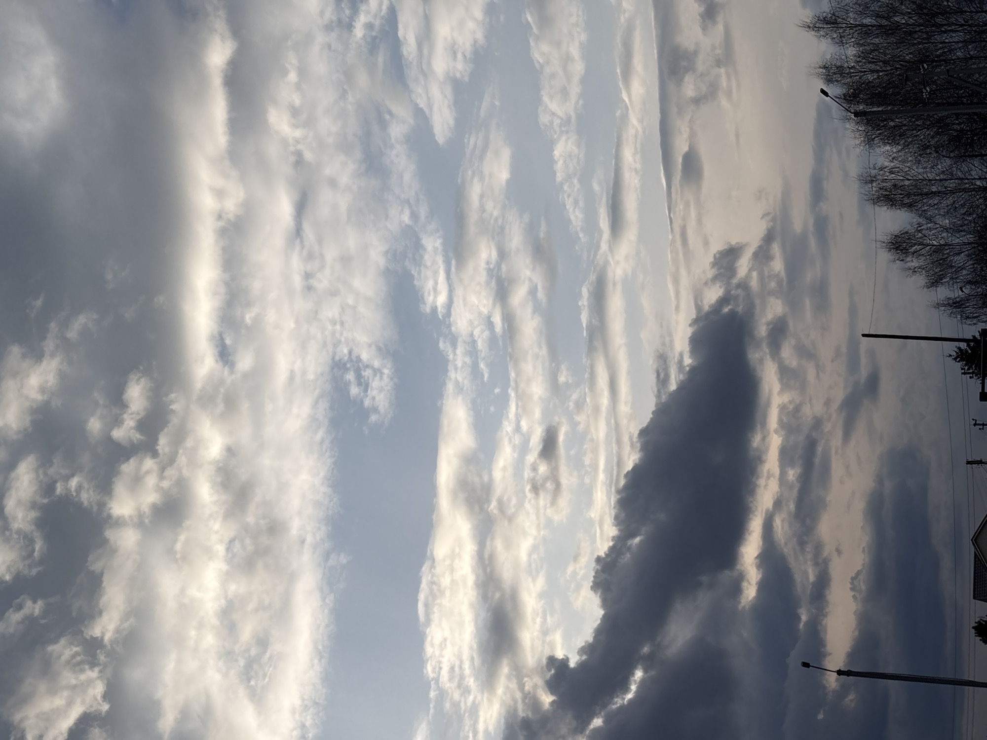 The image shows a view of the sky in Прикамский, Russia. The sky is filled with clouds, ranging in color from white to dark gray. The clouds are arranged in long, vertical formations. On the right side of the image, there are silhouettes of trees and streetlights. There are also some houses that can be seen. The light appears to be soft and diffused.