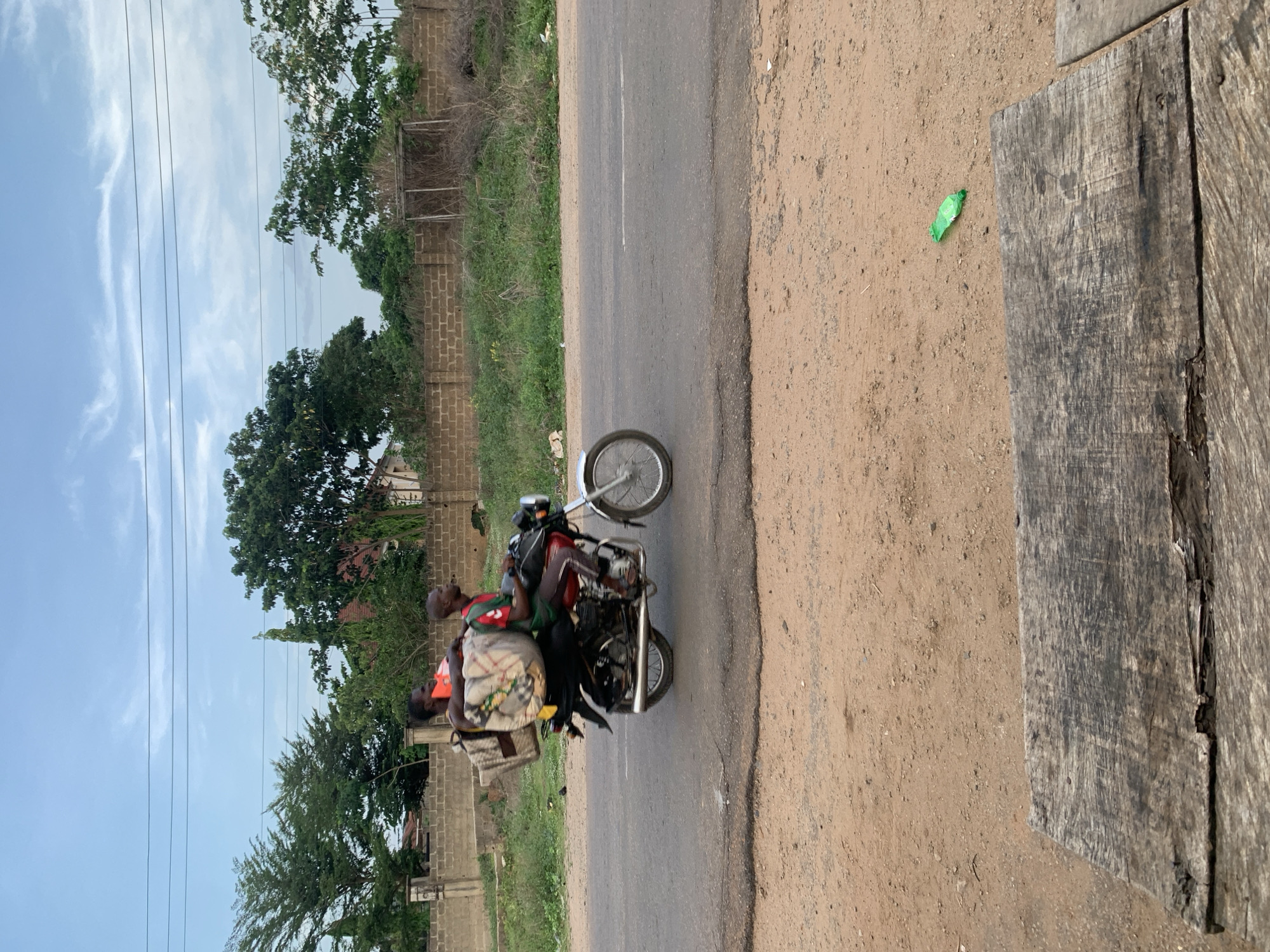 The image depicts a scene in Kabba, Nigeria. A motorcycle with two passengers is traveling along a paved road. The passengers include a driver and a passenger sitting behind him. The passenger is carrying what appears to be a bag or luggage wrapped in a fabric. The roadside consists of a dirt area with some scattered debris, including a green plastic wrapper, and a weathered wooden structure on the right side of the image. On the left of the road is grass, trees, and a brick wall. Overhead power lines are visible in the background against a cloudy sky.