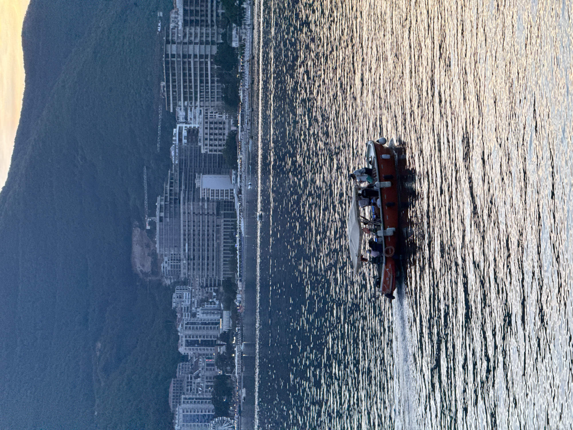 In Budva, Montenegro, on the Adriatic Sea at dusk, a small, red tour boat with a canvas canopy traverses choppy, rippling water. The boat, carrying approximately six passengers, moves from left to right across the frame, leaving a visible wake. The passengers, in casual attire, appear relaxed as they enjoy the evening excursion.
In the background, the coastline is lined with a dense collection of modern, multi-story buildings, characteristic of a developed urban area. Among these structures, construction cranes are visible, indicating ongoing development. The buildings are a mix of light-colored facades and darker glass, with some appearing unfinished. Behind the buildings, a steep, tree-covered mountain rises, its slopes dark and textured. The sky above the mountain is illuminated by the soft, warm hues of the setting sun, casting a gentle glow. The overall scene depicts a lively coastal city during the twilight hours, with the sea activity and urban landscape creating a dynamic contrast.