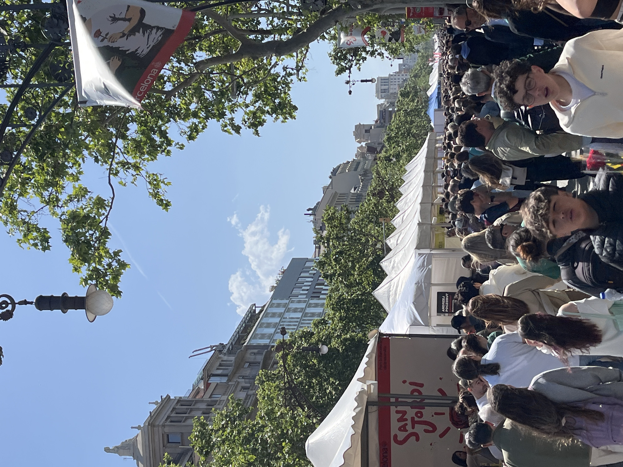 This media file captures a lively outdoor scene, likely a street festival or market. The image is taken from a slightly elevated perspective, looking down a crowded thoroughfare lined with trees and temporary white stalls or tents.
Numerous people are visible, filling the street. Most are adults and young adults, casually dressed, walking in various directions or gathered around the stalls. Some individuals in the foreground are looking towards the camera, while the majority are focused on the event. The atmosphere appears busy and festive.
In the background, multiple-story buildings rise above the tree line, suggesting an urban setting. The sky is clear and blue with a few scattered white clouds, indicating a bright, sunny day. Sunlight casts dappled shadows through the lush green foliage of the trees that line the street. Ornate street lamps are also visible, characteristic of city boulevards.
Several banners and signs are present. A flag-like banner hanging from a lamppost features an illustration of a person holding a rose, with the word "Barcelona" visible at the bottom. Another prominent sign, vertically oriented on a stand next to a tent, clearly displays "Sant Jordi" in large, stylized red letters. This strongly indicates the event is the Sant Jordi festival, a major cultural celebration in Catalonia, Spain, where books and roses are exchanged. On one of the white tents, faintly visible text includes "ANDRZEJ POCZOBUT," possibly a reference or dedication. The overall scene depicts a bustling cultural event in Barcelona on a pleasant day.