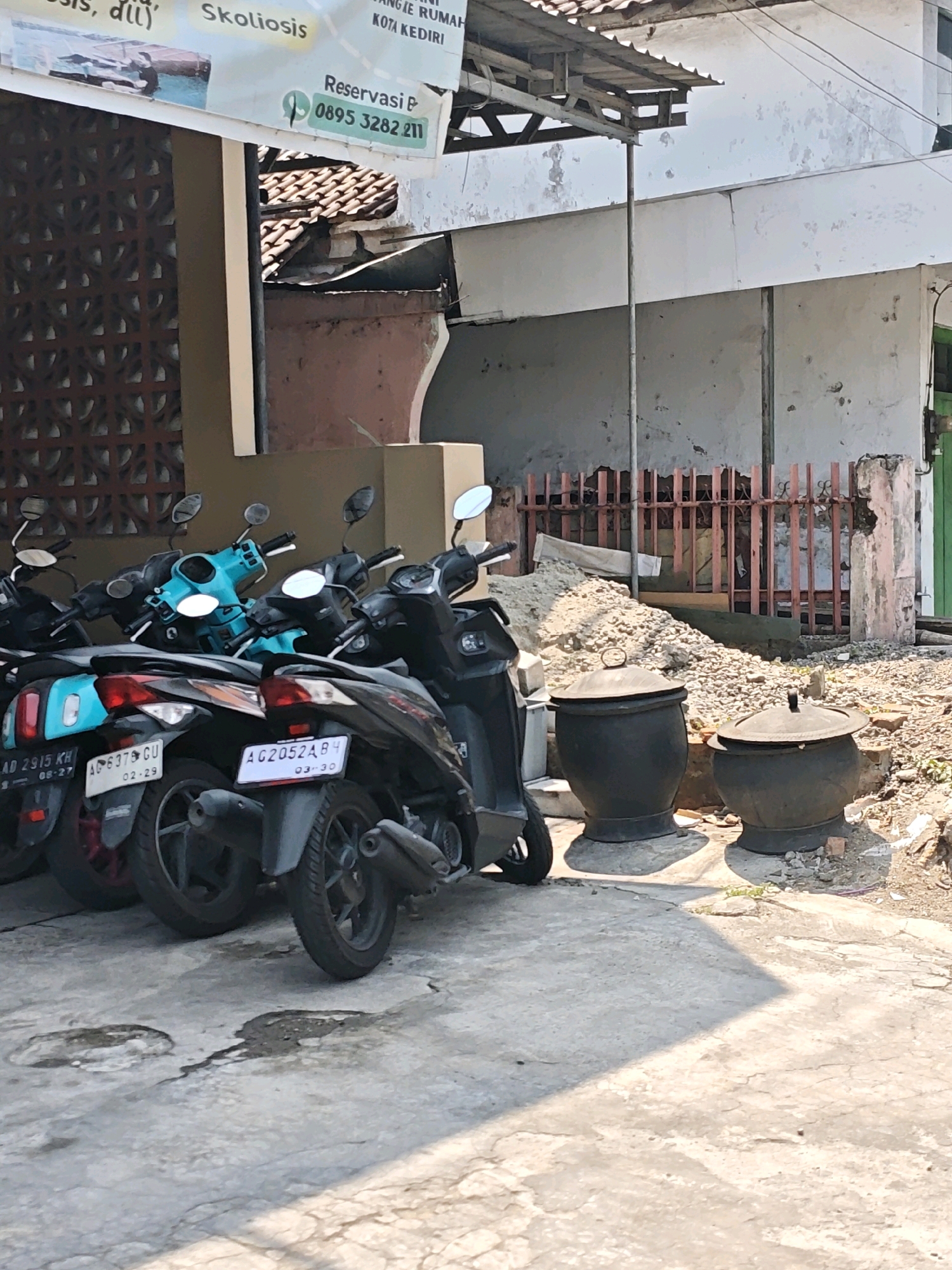 In the city of Kediri, Indonesia, a row of parked motorcycles is visible on a concrete surface in front of a building. The motorcycles, predominantly black and a bright teal, are lined up closely together. One motorcycle has a license plate reading "AG 6379 GU" with the date "02-29". Another license plate is visible, "AG2052ABH4" with the date "03-30". In the background, two large, dark, lidded pots stand on the ground, surrounded by a pile of gravel and debris. A weathered, reddish-brown fence with vertical slats is situated behind the pots. The building appears to be under renovation or in disrepair, with cracked and stained walls. A sign with text is partially visible at the top left of the image, including the words "Skoliosis" and a phone number "0895 3282 211". The scene suggests an outdoor area in a commercial or residential district of the city, with evidence of recent construction or maintenance activities.