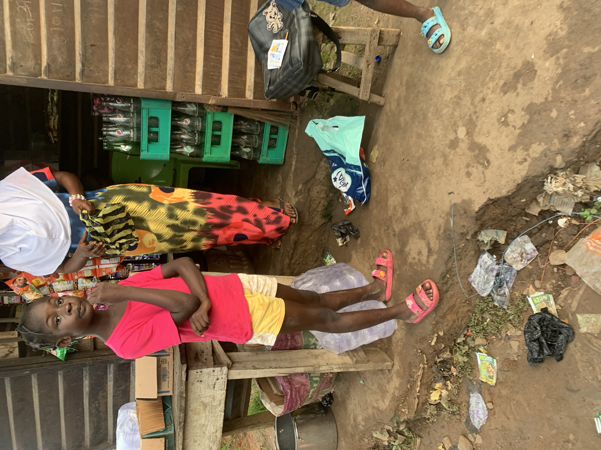 A young Black girl stands at a roadside stand in Shagamu, Nigeria, in front of a wooden stall. She wears a bright pink t-shirt and yellow and white patterned shorts, along with pink slide sandals. Her arms are crossed, and she holds her chin. To her right, partially visible, an adult woman wears a white headscarf and a colorful, patterned dress. Behind the girl, on a wooden shelf, are boxes, and to the right, green crates hold glass bottles of Pepsi. The ground is dirt and scattered with litter. A black bag rests on a wooden stool to the right of the scene.