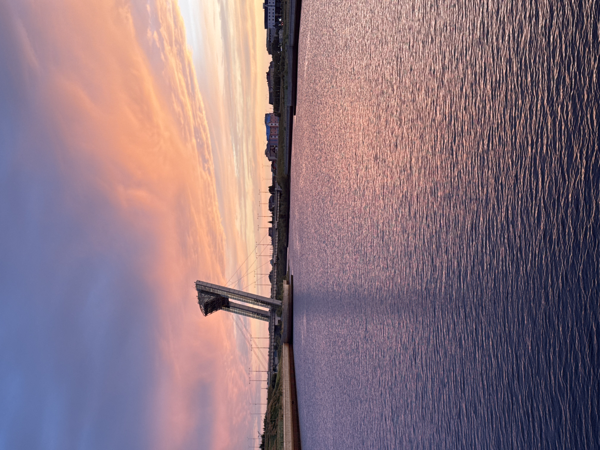 This image captures a sunset over the Mamonov Canal in Salekhard, Russia. A distinctive cable-stayed bridge, part of the Salekhard-Igarka Railway, dominates the left side of the frame, its angular structure silhouetted against the vibrant sky. The sky itself is a gradient of soft blues and fiery oranges, with wispy clouds catching the last rays of the sun. The canal's surface reflects the colorful sky, creating a serene and tranquil atmosphere. In the distance, a row of buildings forms the city skyline, sparsely lit by streetlights. The foreground is filled with the rippling water of the canal, its surface a deep blue-purple hue, subtly reflecting the pinks and oranges of the sunset. There are no people visible in the image, and the scene depicts a still, quiet urban landscape at dusk.