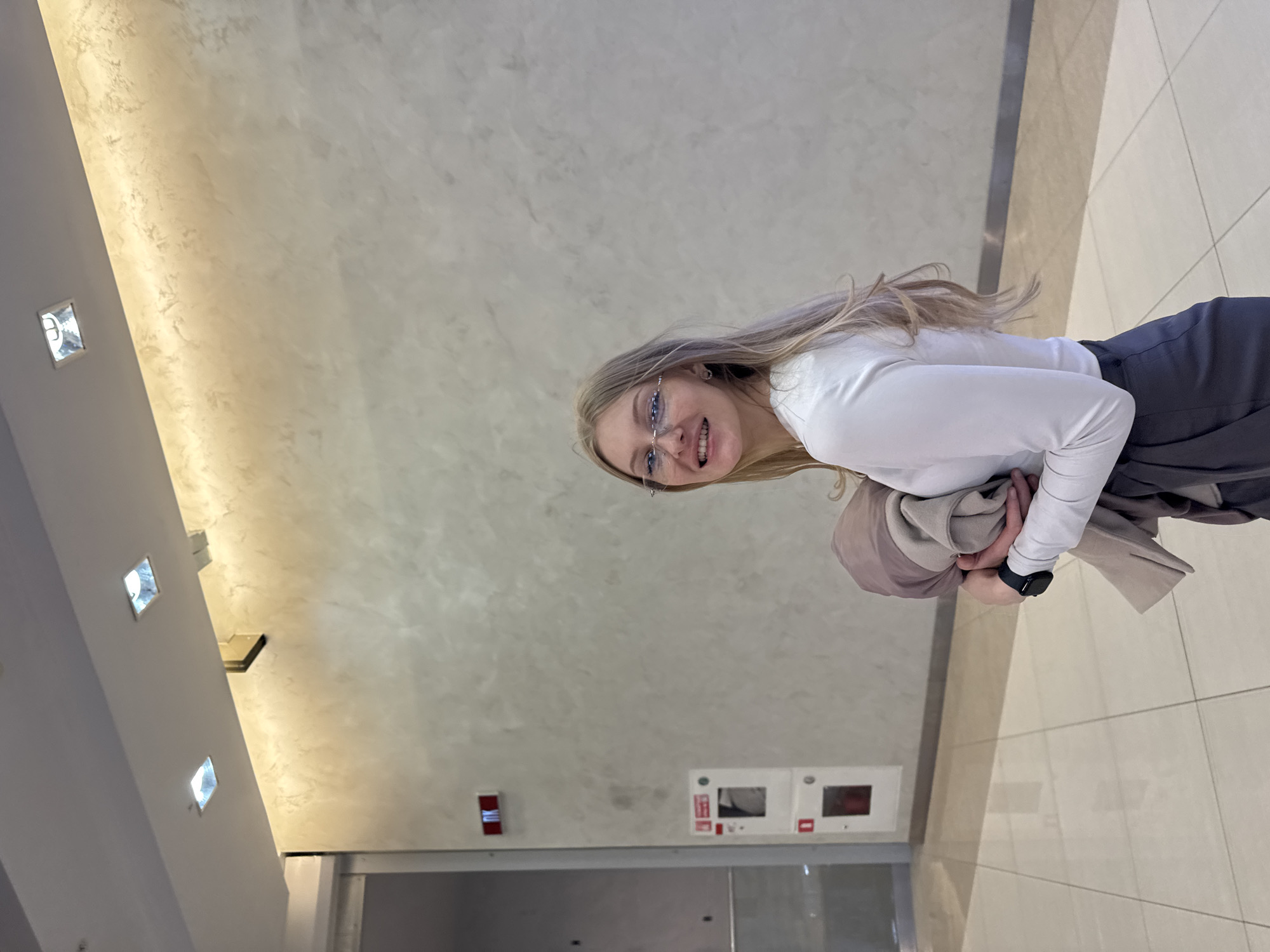 A young woman with long blonde hair, wearing glasses, a white long-sleeved shirt, and gray trousers, stands in what appears to be an interior space with a light-colored, textured wall and tiled floor. She smiles at the camera while holding a beige jacket or coat.
The wall features recessed lighting and several rectangular panels with red and white elements near the bottom. A metal doorframe suggests the entrance to a room or elevator.
The location appears to be an interior space in Perm, Russia, based on the architectural details and visual cues.