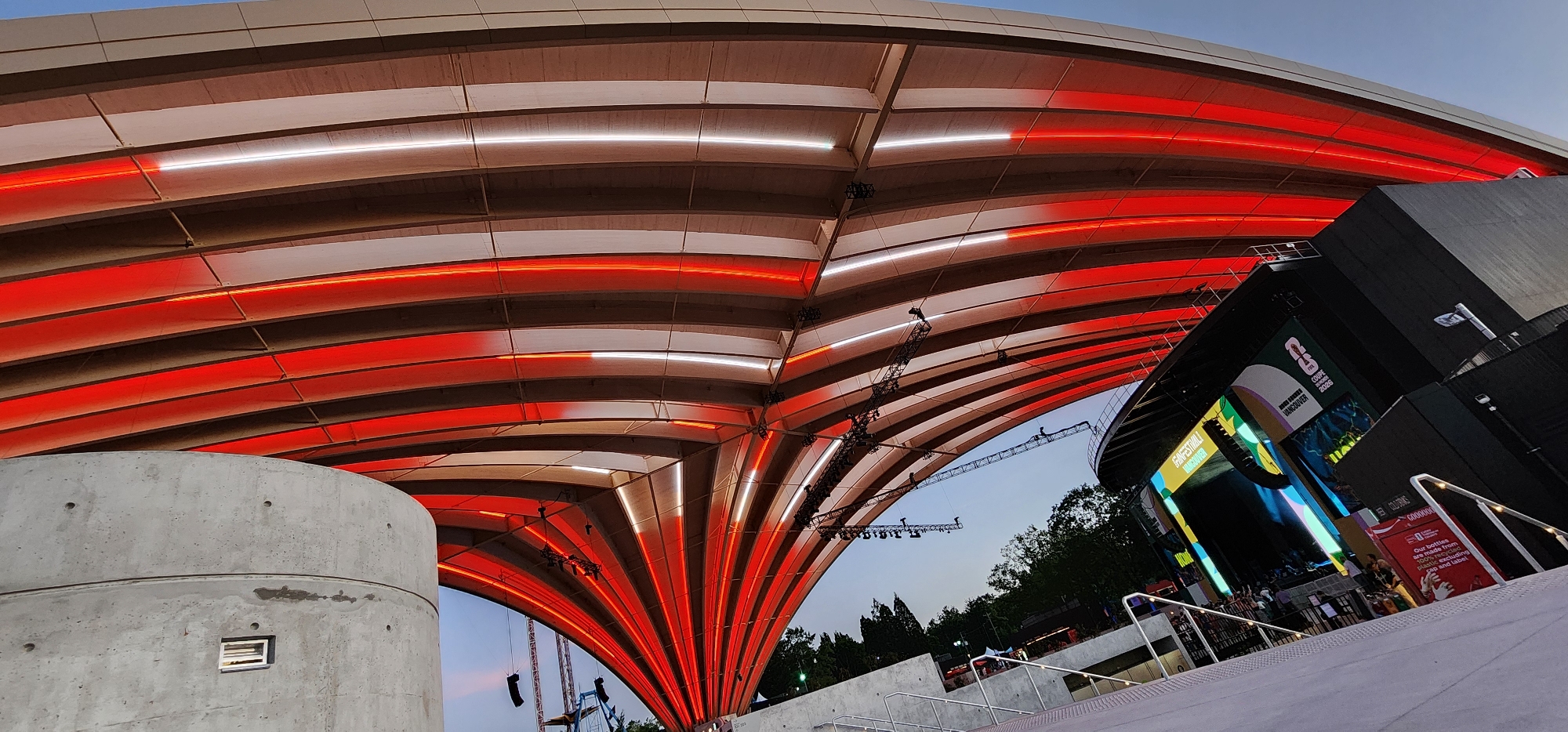 An exterior, low-angle, wide shot captures a modern outdoor venue with a large, curved, slatted roof structure illuminated with alternating red and white lights. The roof's wooden slats are arranged in parallel lines, curving upwards and outwards from a central point. Below the roof, a stage is visible on the right side of the image, featuring a large screen displaying graphics and text, including the words "FANFESTIVAL VANCOUVER" and "COupe 2026". A black, angular building serves as the backdrop for the stage. In the foreground, a curved, grey concrete structure with a small, rectangular opening is visible on the left. Stairs and railings lead up to the viewing area in front of the stage. Trees and a clear sky are visible in the background. Several pieces of lighting and sound equipment are suspended from the roof structure. A few distant figures are visible on the stage area.