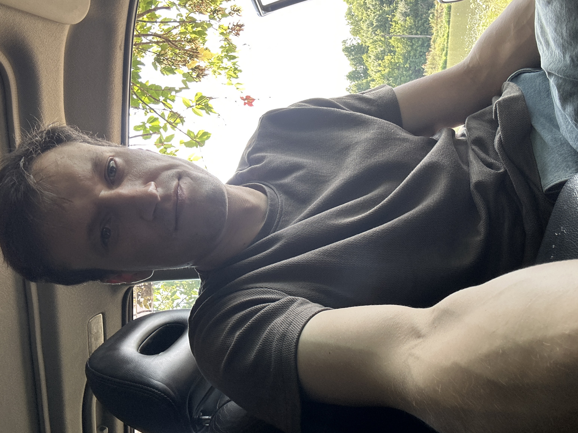 A young Caucasian man with short brown hair and stubble is seen from the chest up, sitting in the driver's seat of a vehicle, looking at the camera. He is wearing a dark grey short-sleeved polo shirt and blue jeans. The interior of the car is visible behind him, including a black headrest. Through the car window to the right, a body of water, lush green trees, and a bright sky are visible, suggesting an outdoor, possibly rural, setting outside of Makiv, Ukraine. The lighting indicates it is daytime, likely late morning or early afternoon.