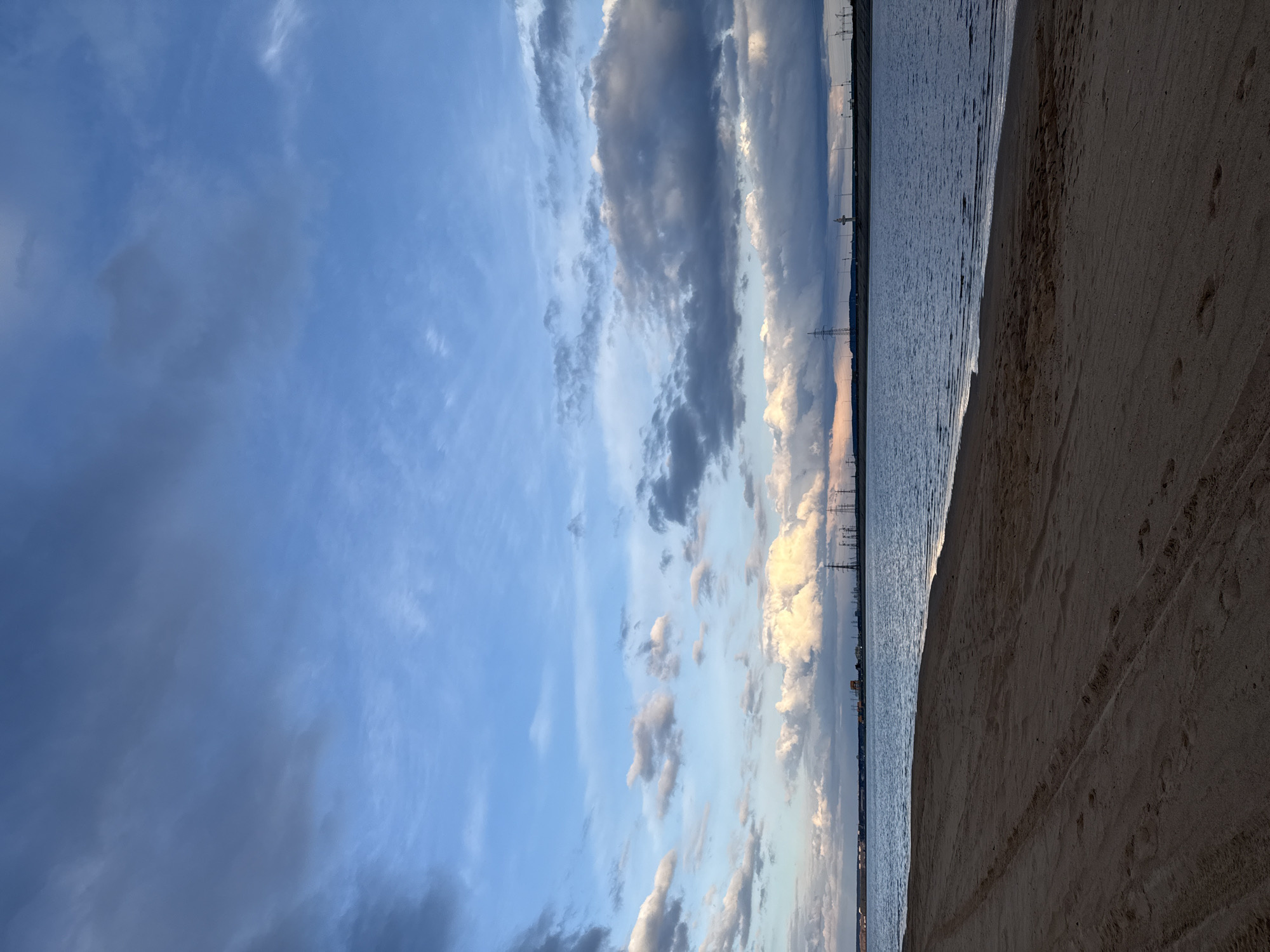 This image depicts a serene coastal landscape in Noviy, Russia, likely captured during the late afternoon or early evening. The scene is composed of a vast sky, a body of water, and a sandy beach.
The sky occupies the upper two-thirds of the frame, showcasing a mix of light blue hues and dramatic cloud formations. Some clouds are bright white and illuminated by the soft, warm light of the setting sun, while others are darker grey and more elongated, creating a sense of depth and movement.
Below the sky, a calm body of water, likely a sea or a large bay, extends across the mid-ground. Its surface appears relatively still, with subtle ripples visible near the shoreline. Along the distant horizon, faint silhouettes of industrial structures, specifically power lines or transmission towers, are discernible, indicating the proximity of developed land.
The lower third of the image features a wide, sandy beach. The sand appears damp and darker closer to the water's edge, gradually becoming lighter further inland. Visible on the sand are numerous footprints and distinct tire or tread marks, suggesting recent activity by individuals and possibly small vehicles.
No people or specific activities are taking place; the scene conveys a peaceful and expansive mood. There is no visible text in the image.