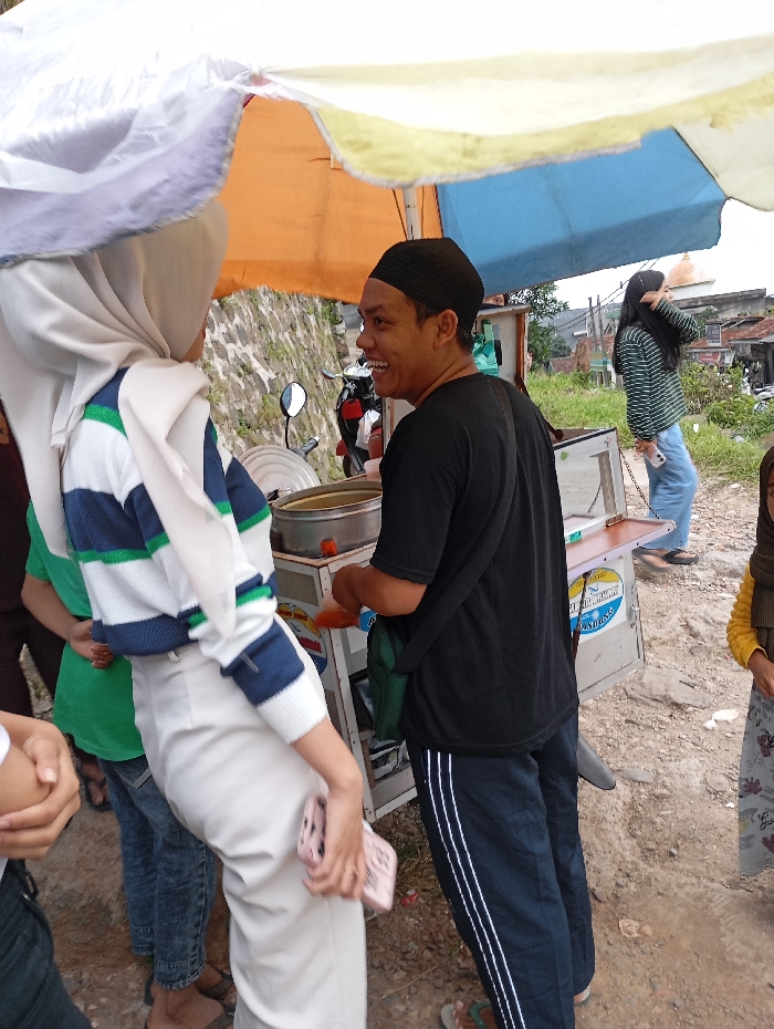 The scene takes place on a street in the Kecamatan Kutawaringin district of Indonesia. A man with a black skullcap is standing next to a food cart and talking to a woman wearing a white hijab and white pants. The man is holding a green bag slung across his shoulder and the woman is holding a pink phone in her hand. The woman has a white hijab wrapped around her head. There are other people standing nearby, including a woman with a grey shirt and blue pants and a young girl in a yellow and pink shirt and grey pants. The food cart is surrounded by a white canopy and has a sign on the side that reads "Mie Aceh". The ground is paved and there are a few motorcycles parked in the background. The weather is clear and the sky is overcast.