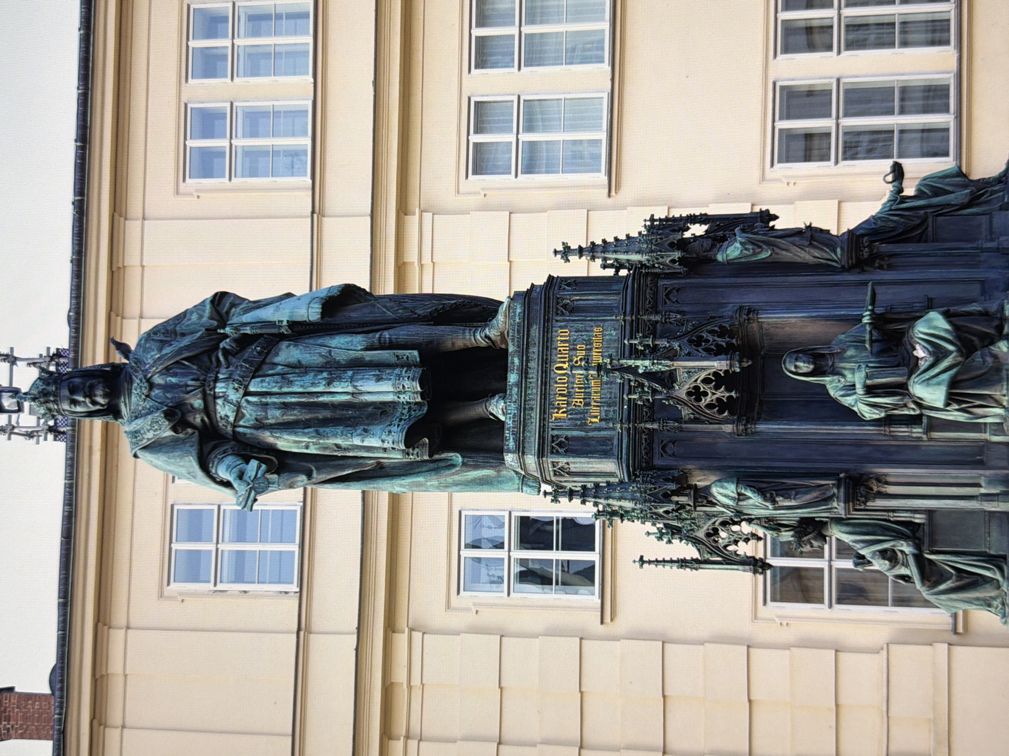 The image shows a statue of a king with a crown, holding a scepter in his right hand and a scroll in his left hand. The statue is made of bronze and is a weathered greenish-blue color. The king is wearing a long robe and a crown. He is standing on a large pedestal which is decorated with ornate gothic architecture and smaller statues. There is an inscription on the pedestal. The statue is located in front of a building with a light-colored facade and several windows. The building has a classical architectural style. The statue is detailed and appears to be a historical monument. The background shows the building and the sky. There are no people visible in the image other than those depicted in the statue. The setting appears to be a city square or a courtyard. The statue is the central focus of the image.