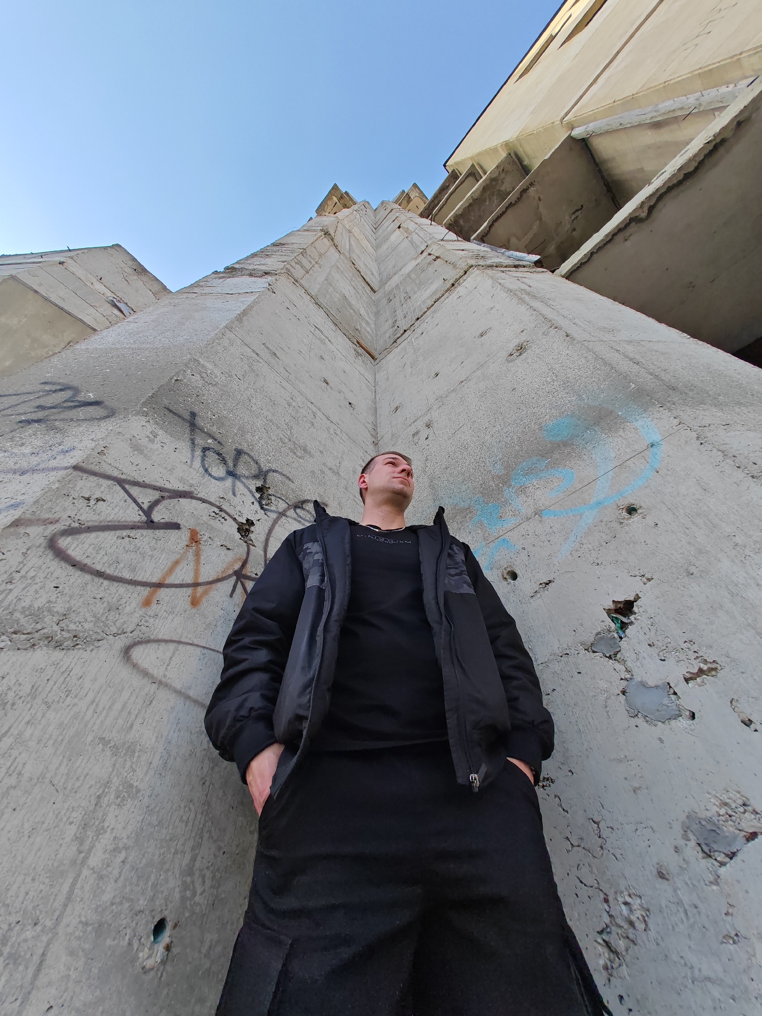 An upward-angled photograph depicts a male figure standing against the base of a massive, angular concrete structure. The scene is located at the Monument to 1300 Years of Bulgaria (Founders' Monument) in Shumen, Bulgaria, specifically within its complex of monumental concrete forms.
The subject is a male, appearing to be in his 20s or 30s, with short hair. He is dressed entirely in black, wearing a black t-shirt, a black puffer jacket, and black pants. His hands are placed in his jacket pockets. He stands with his head tilted slightly upward and to the viewer's left, looking in that direction with a neutral expression.
Behind the subject, the concrete wall exhibits coarse texture, minor damage, and multiple instances of graffiti. Visible graffiti includes dark blue, light blue, and orange-brown spray paint forming indistinct letters and abstract shapes. The concrete itself shows exposed aggregate and signs of weathering and minor spalling.
Above the subject and extending vertically, the immense concrete structure ascends sharply, characterized by repetitive, jutting angular planes and deep shadows. The upper portions of the structure appear unfinished or degraded, with exposed rebar elements and rough concrete surfaces. The background reveals a clear, light blue sky, indicating daylight conditions. The overall environment presents a stark, industrial aesthetic.