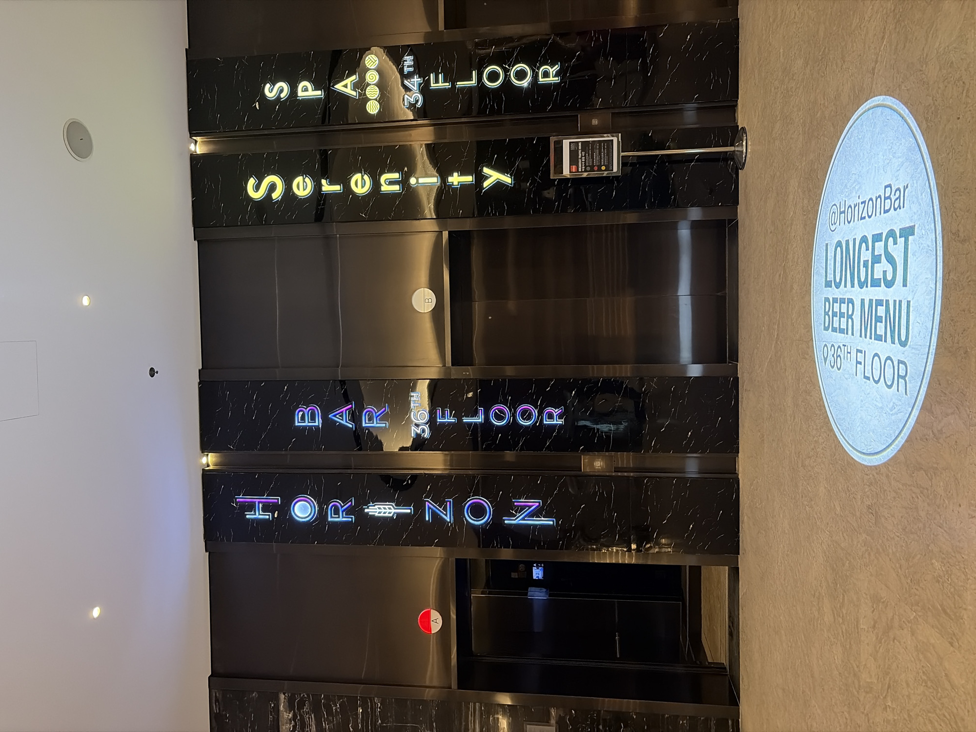 The image shows an interior scene of a building lobby with multiple elevators. The elevators are flanked by vertical, illuminated signs. The leftmost sign spells out "HORIZON" in multicolored neon letters, with an icon resembling a wheat stalk between "R" and "I", and a symbol for floor "A" is visible on the elevator door to its left. The sign to the right of "HORIZON" spells out "BAR" in multicolored neon letters, followed by "36th FLOOR" in white neon. Further to the right, a sign spells "Serenity" in yellow neon letters. The rightmost sign spells "SPA" in green neon letters, followed by "34th FLOOR" in white neon. A floor projection in the foreground displays an oval shape with text that reads "LONGEST BEER MENU 36TH FLOOR" and "@Horizonbar". A sign on a stand is positioned in front of the rightmost elevator. The overall environment appears to be a modern and upscale building. The scene is set in Đà Nẵng, Vietnam.