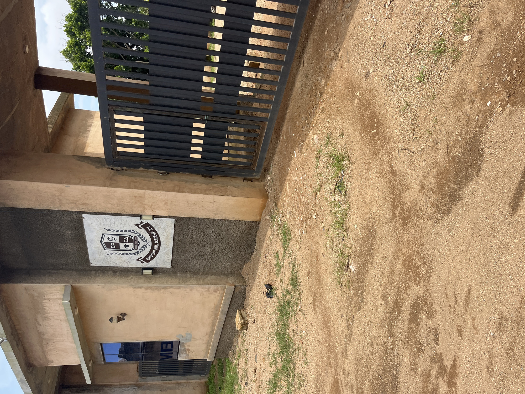 The image shows the entrance to Citipoint Intl. Academy, JOS, located in Jos, Nigeria. A dark metal gate is partially open, revealing a glimpse of the compound beyond. To the left of the gate, on the wall of the building, is the school's crest, which features the initials "CP EA" within a shield. The building itself appears to be constructed with rough, unfinished concrete and painted in a light, earthy tone. A small window with blue-tinted glass is visible to the left of the crest, and a dark metal grill is in front of it. The ground in front of the academy is covered in loose sand and dirt, with sparse patches of dry grass. A single rock sits on the ground near the base of the building. The overall impression is one of a functional, but not elaborately decorated, educational institution.