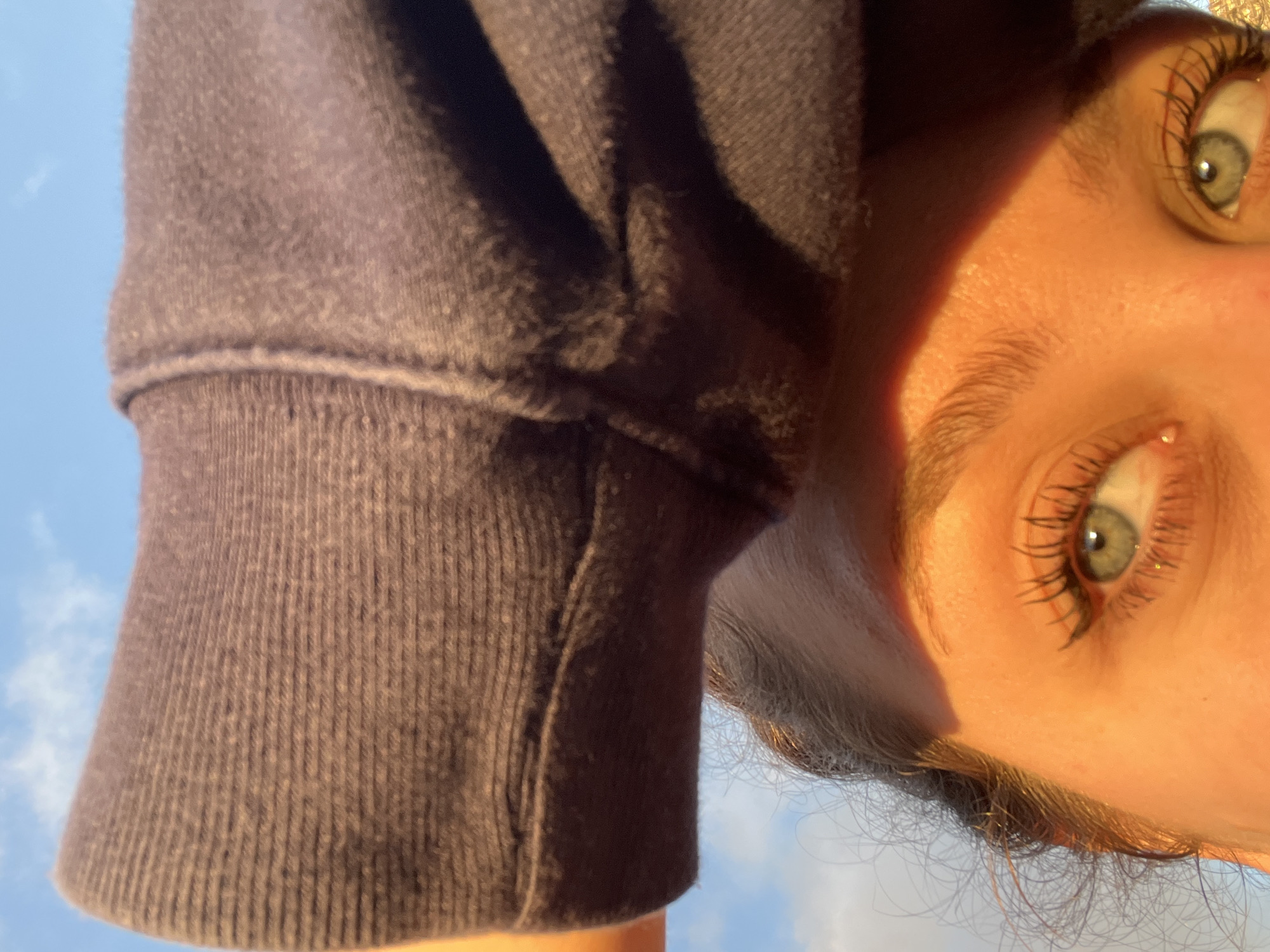 The image shows a person's face at an unusual angle, with the sky visible in the background. The person has fair skin, light-colored eyes, and visible eyelashes. A portion of a gray-purple sweatshirt is also visible, suggesting the person is wearing it. The photo appears to be taken outdoors, possibly in the daytime. There is no information to suggest that this photograph was taken in Mogán, Spain.
