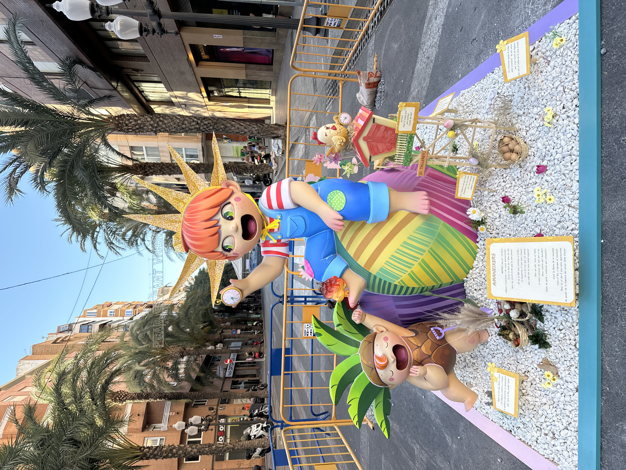 This is a vertical, medium shot of a large, colorful, street art display, likely a "falla" from the Valencian Community, Spain. The subjects are two cartoonish figures, one larger with orange hair and a yellow sunburst headdress, wearing a blue outfit with red and white striped sleeves, and holding a pocket watch. The other figure is smaller, lying on a bed of white pebbles, with brown hair and a shell-like headdress, wearing a brown outfit. The display is on a street, with buildings and palm trees in the background. There are also decorative elements like a small wooden structure with Easter eggs and flowers, and small signs with text in Spanish and Catalan. The scene appears to be set up outdoors during the daytime, with clear blue skies visible. The overall mood is festive and playful, indicated by the exaggerated expressions of the figures and the vibrant colors of the display. Signs visible include "FCARNER SANT VICEN", "EL TALLERET", and some handwritten text in Spanish and Catalan.