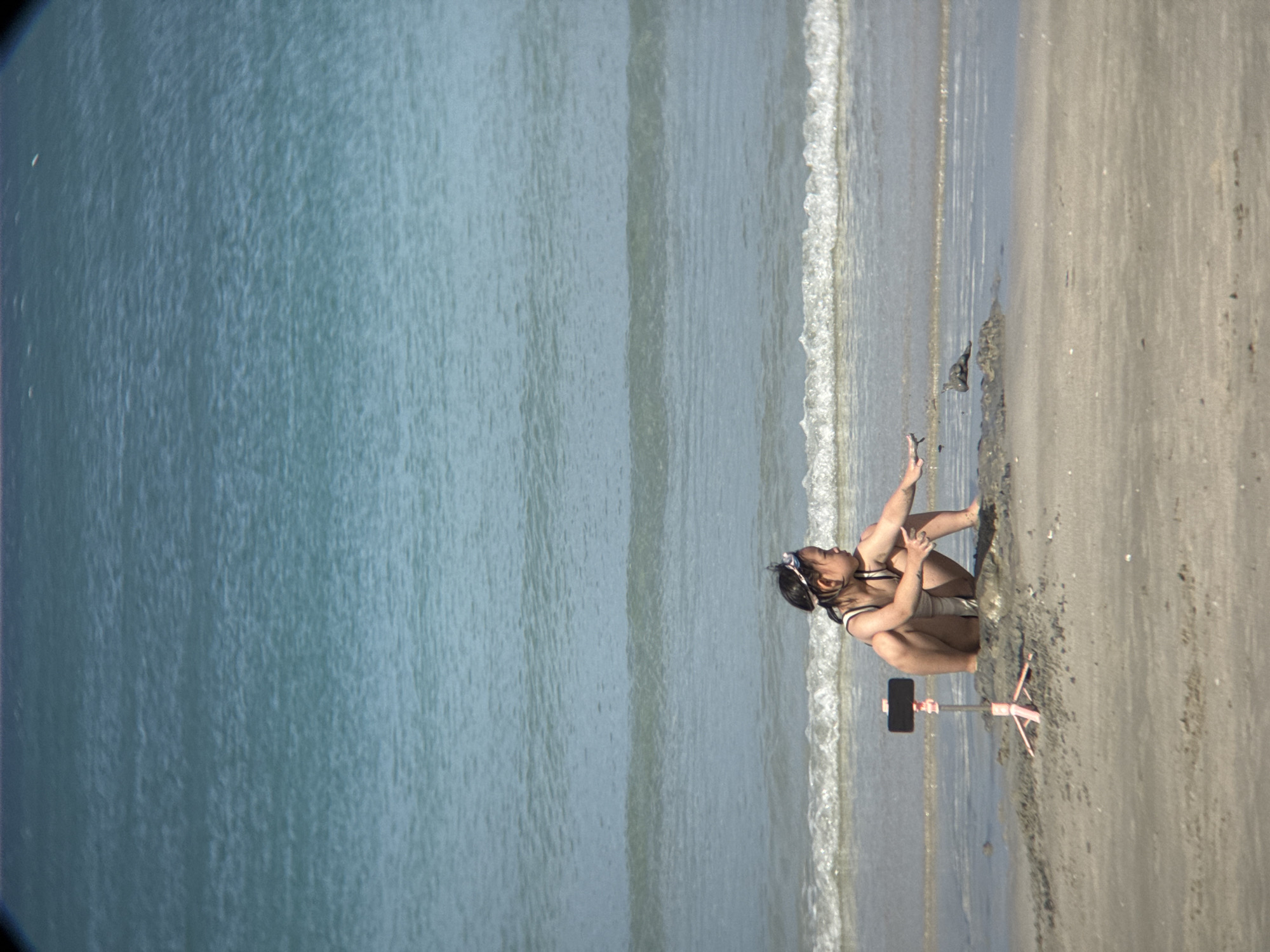 This is a photograph taken at a beach in Đông Ninh Hòa, Vietnam. The image features a young woman in a white swimsuit crouching on the wet sand near the water's edge. She is looking towards a smartphone mounted on a small tripod, suggesting she might be recording a video or taking a selfie. Her body is positioned to face the camera, and her right arm is extended upwards, with her hand slightly open. She is wearing a headband, possibly to keep her hair out of her face.
The scene depicts a bright, sunny day. The ocean on the left side of the frame is a calm, blue expanse with gentle waves lapping onto the shore. The sand on the right is a light brown color, with some dampness from the receding tide. The water creates a clear line where it meets the sand, with a foamy white edge from the breaking waves. The overall mood of the image appears relaxed and peaceful, with the woman enjoying her time at the beach. There is no visible text in the image.