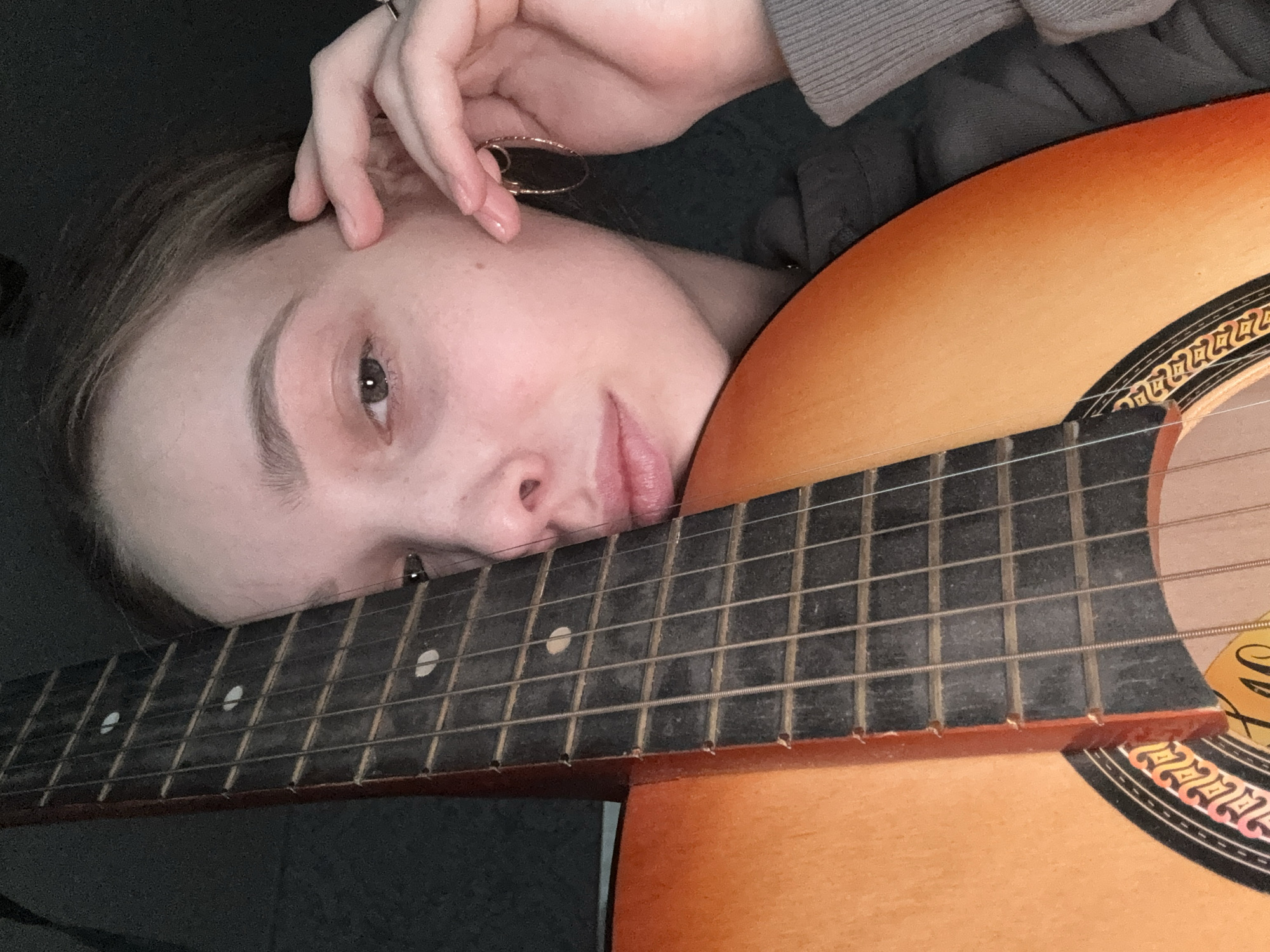 A young person is resting their head on a guitar. The guitar has a dark fretboard with light inlays, and a reddish-orange body. The person is facing the camera. There is nothing in the image that indicates it was taken in Прикамский, Russia.