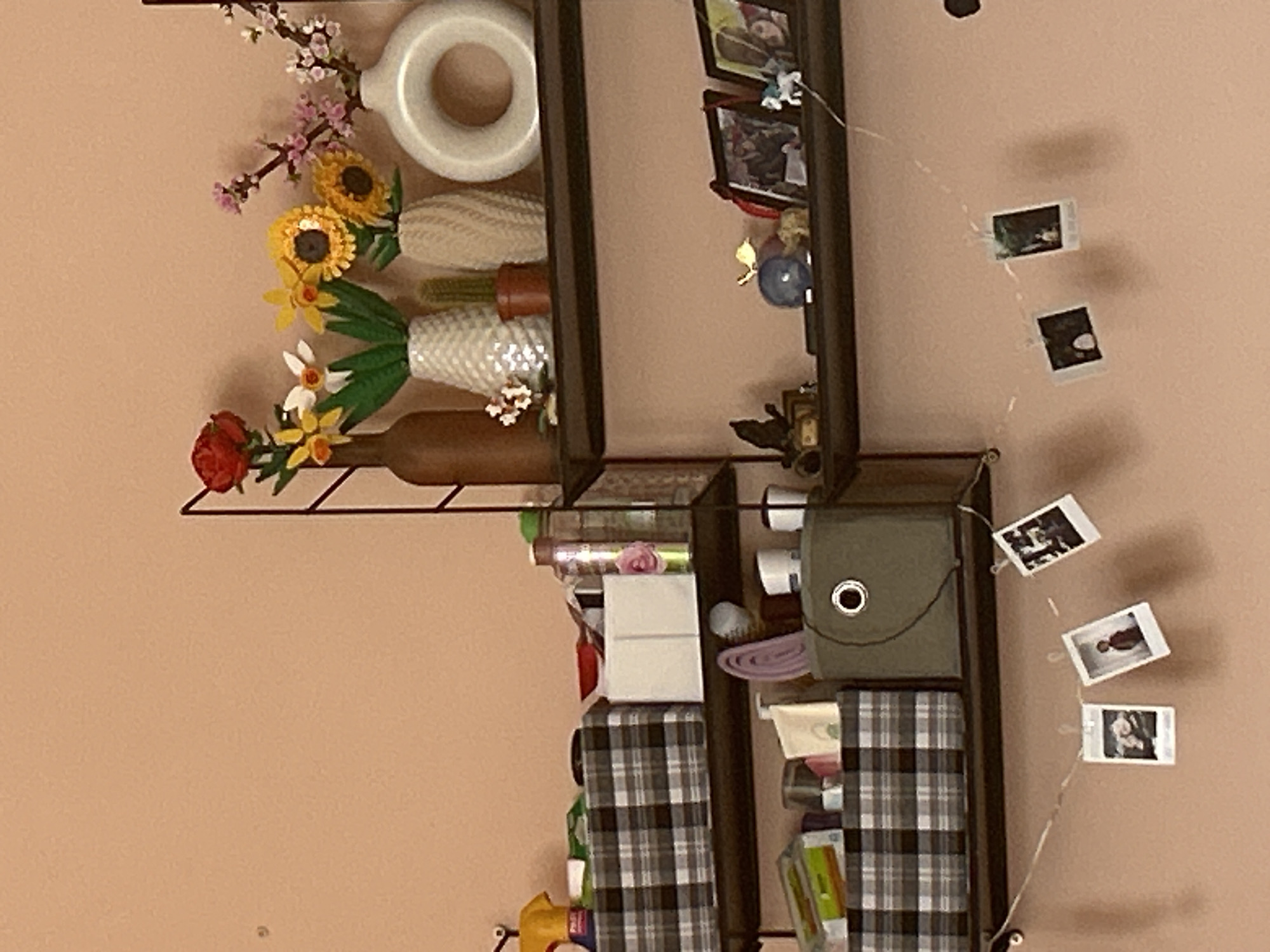 The image shows a vertically oriented wall with two dark brown shelving units attached. The wall is painted a pale peach color.
The left shelf contains decorative objects: a white ceramic donut-shaped object, faux flowers (including sunflowers, daffodils, and cherry blossoms), and a brown glass bottle.
The right shelf contains a variety of items including framed photos, a small toy figurine, a green canvas bag, and various toiletry or household items in boxes and containers. The lower shelves have gray and white plaid fabric-covered boxes.
Strands of small photographs are clipped to the wall, running diagonally down and to the right from the upper right shelf.
There is no information in the picture that reveals the location as Sofia, Bulgaria.
