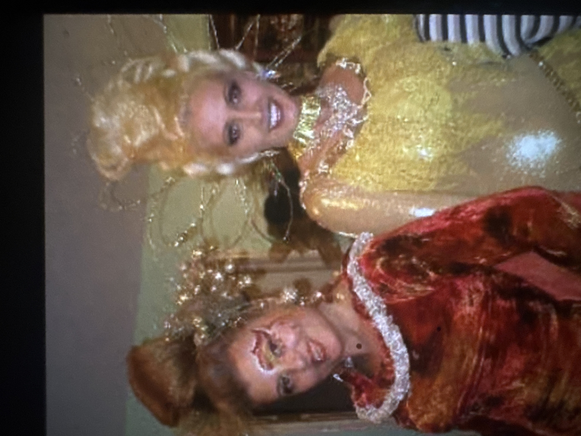 The image displays two women in elaborate costumes, posing for a photograph indoors. The original image is rotated ninety degrees counter-clockwise.
Viewing the image upright, the woman on the left features styled blonde hair with sparkling accessories and a bright smile directed at the camera. She wears a vibrant yellow or gold sequined dress adorned with a ruffled, white and gold collar.
The woman on the right has reddish-brown hair, also styled elaborately with sparkling embellishments. She is dressed in a rich red or burgundy gown with a similar white ruffled collar. Her face exhibits prominent special effects makeup, notably around her left eye and cheek, giving the appearance of a facial injury or theatrical wound. Her expression is more neutral as she also looks towards the camera.
Both women's attire suggests a costume party, masquerade, or a themed event. The background is softly blurred, indicating an indoor setting. There is no visible text in the image. The lighting appears to be artificial.