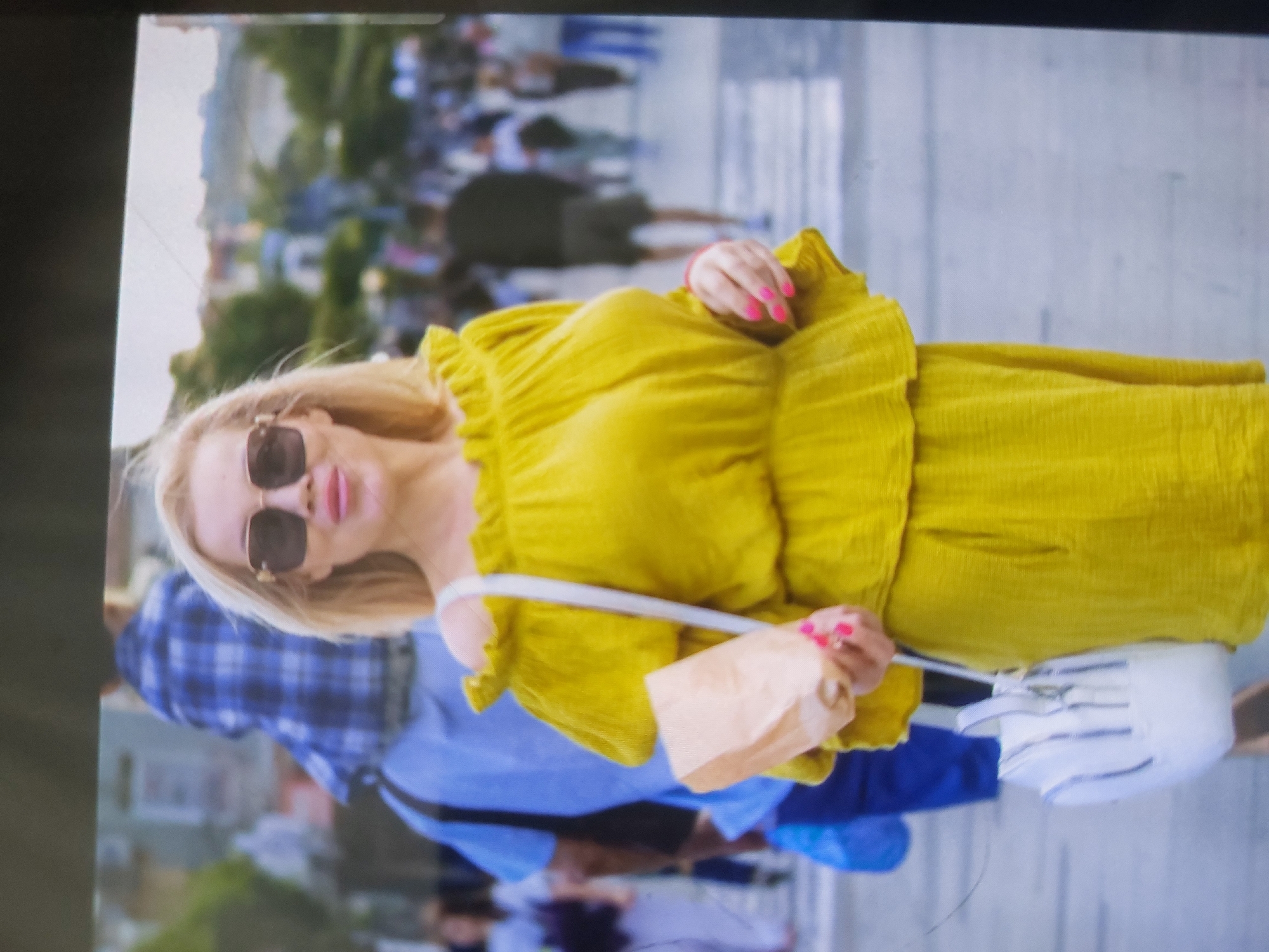 A photograph depicts a blonde woman wearing a yellow dress and sunglasses. She is carrying a white purse and a brown paper bag. The background shows a crowd of people on a city street. The exact location in Tsarevo, Bulgaria, is not discernible from the image.