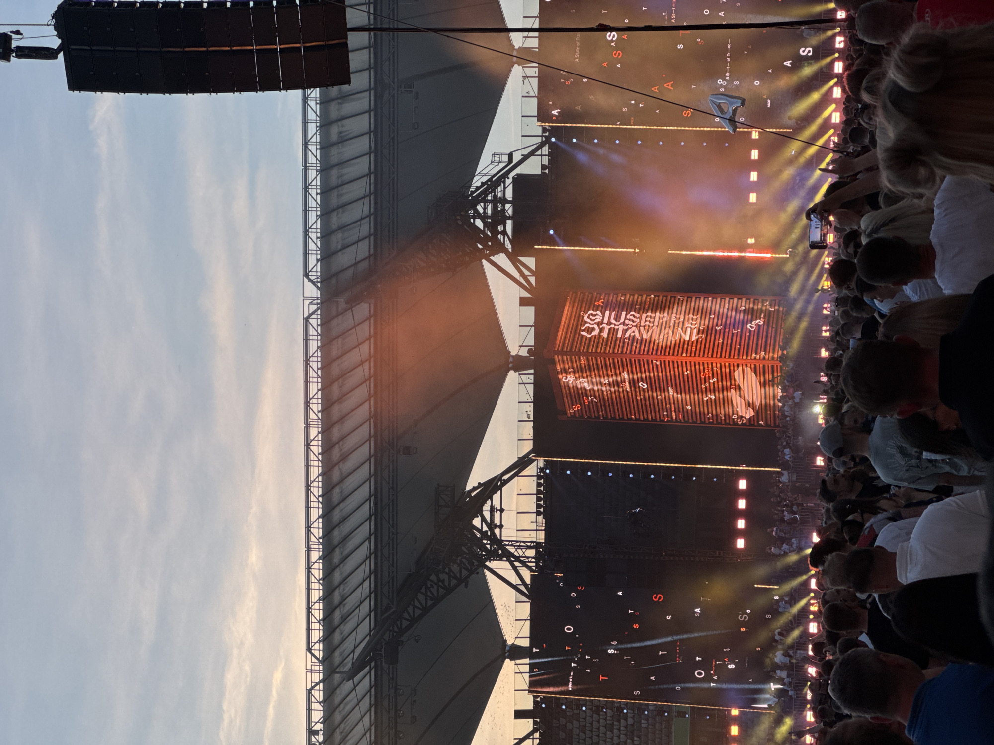 The image captures an outdoor concert at the PGE Narodowy stadium in Warsaw, Poland. A large crowd of people stands in front of a massive stage adorned with illuminated screens and lighting rigs. A central screen displays a vertical, orange, corrugated structure with white text. To the left and right of this central display are large LED screens showing abstract patterns of letters and symbols, illuminated by stage lights. The stadium's roof structure is visible in the background against a twilight sky. Several large speaker arrays are suspended from above, one prominently positioned on the right side of the frame. The atmosphere is lively, with many attendees holding up their phones to record the event.
