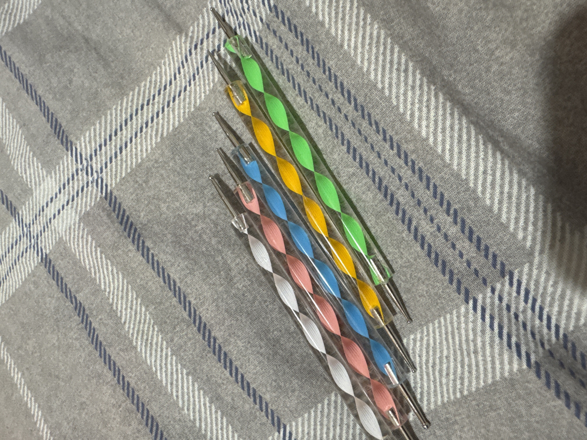 Five dotting tools with colorful spiral handles are laid out diagonally on a grey and white plaid fabric. The tools have silver metal tips. From left to right, the handles are white, pink, blue, yellow, and green. The fabric appears to be a blanket or bedspread. There is no indication of the location being in the city of Яромінскі сельскі Савет, Belarus.